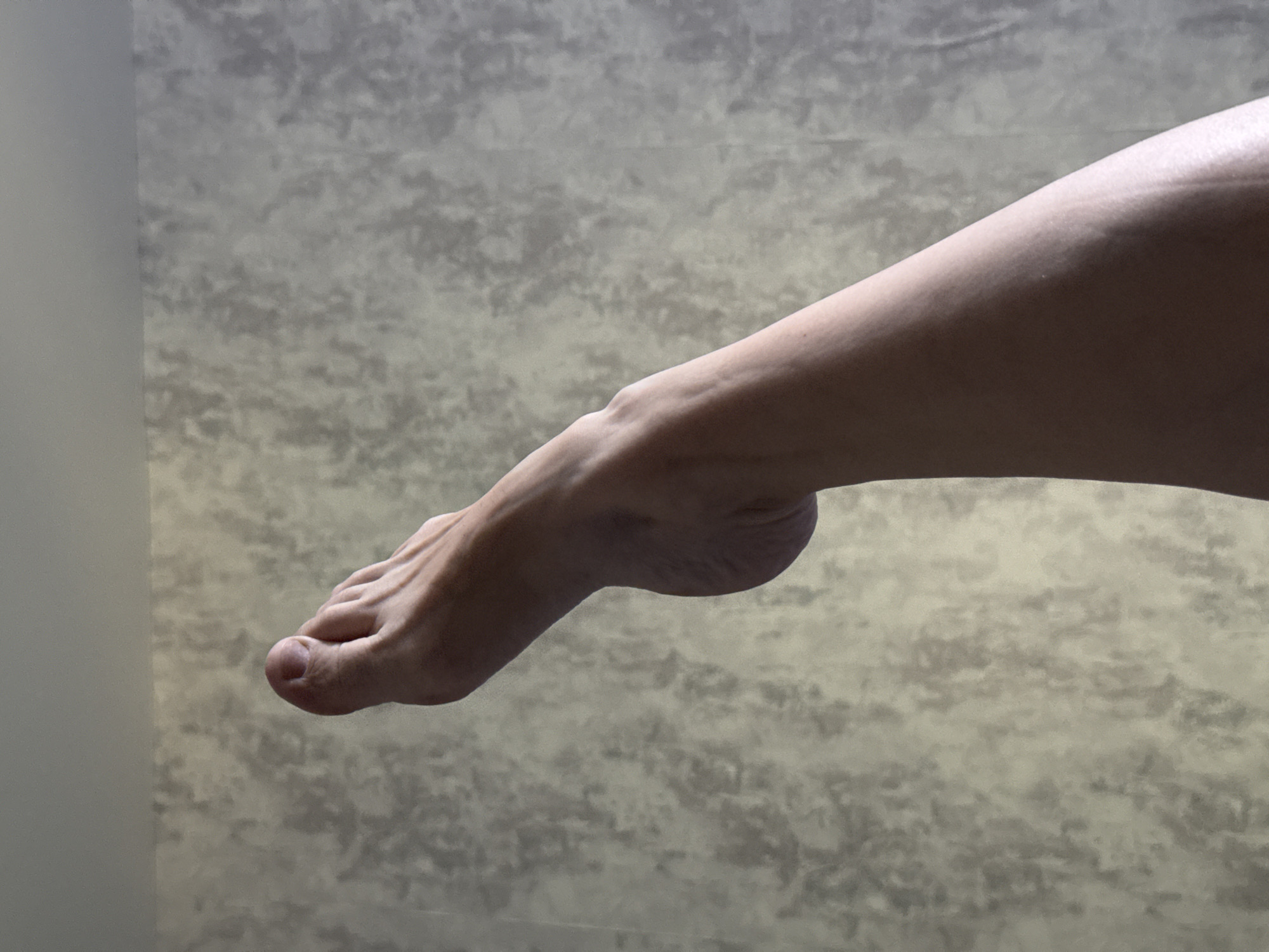 A close-up, high-angle shot shows a person's leg and foot extended upwards against a textured, neutral-colored wall. The foot is bare, with toes pointed, and the leg appears smooth. The wall has a mottled pattern, with lighter and darker grey and beige tones. The lighting casts soft shadows, highlighting the contours of the leg and foot. No specific location in Yekaterinburg, Russia can be identified from the image.