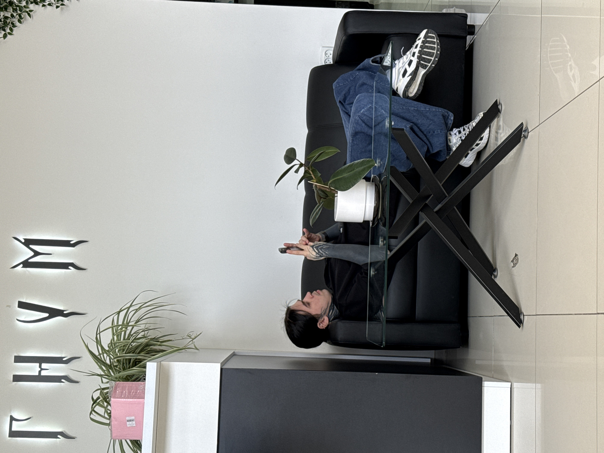 In an indoor setting, a person is seen lying comfortably on a black leather couch, head to the left and feet propped up on the backrest. They are wearing blue jeans and white sneakers with black accents, and a dark long-sleeved top that reveals extensive black tattoos on their arms and hands. The person is looking down at a smartphone held in both hands, appearing relaxed and engrossed.
In front of the couch, there is a modern glass-topped coffee table with a distinctive black X-shaped base. A small potted plant with dark green leaves sits on the table. To the left, a white and black counter or cabinet structure is visible, hosting another potted plant with long, thin variegated leaves, and a pink packaged item. The walls are plain white, with a series of stylized, illuminated black letters mounted on the wall to the left, which spell out "МАЯК" (MAYAK). Light-colored tiled flooring extends throughout the space. Electrical outlets are visible on the white wall behind the couch. The overall ambiance suggests a clean, contemporary commercial or waiting area, possibly a salon or studio, consistent with the location context of Rostov-on-Don, Russia.