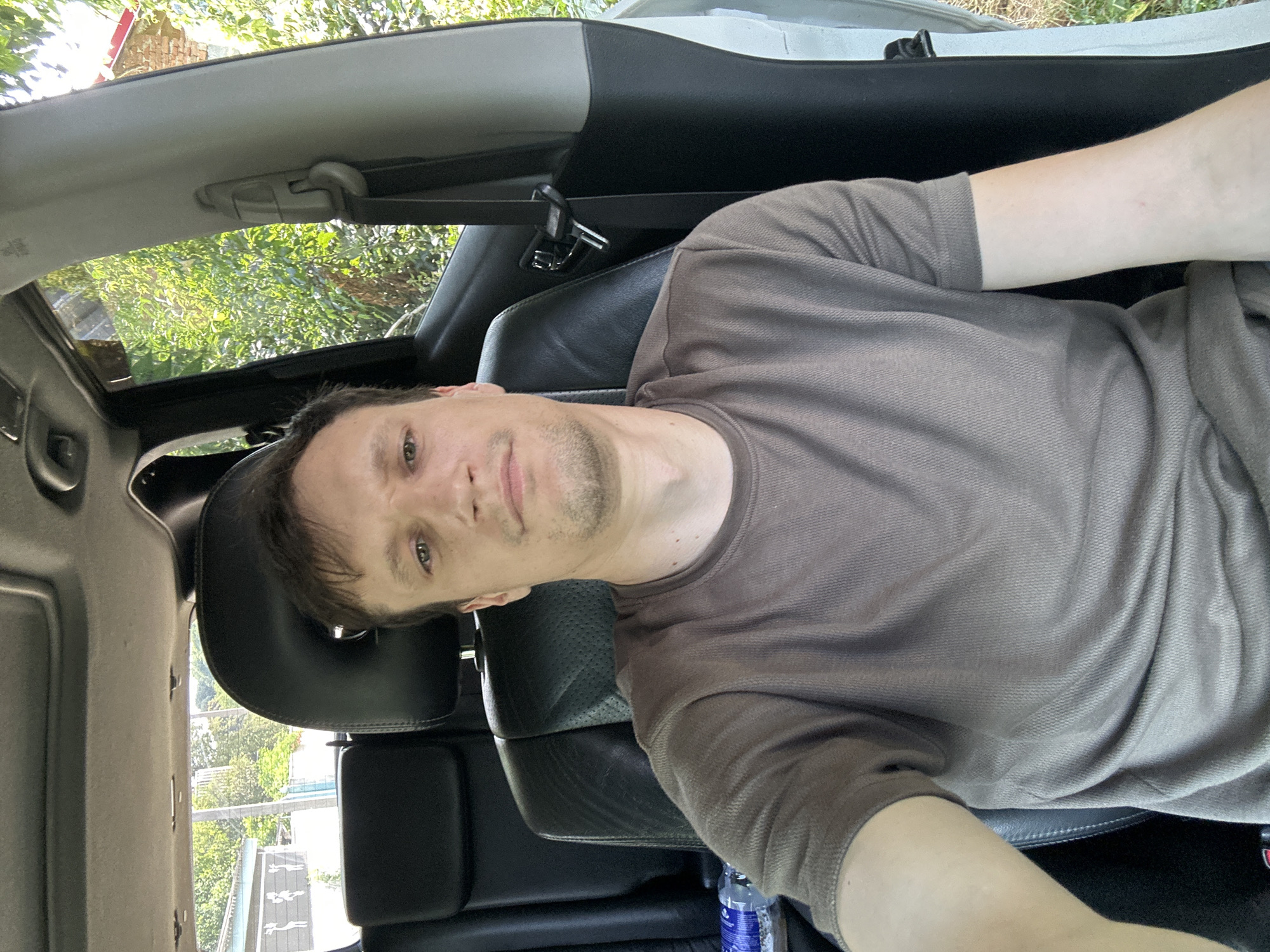 A man is sitting in the driver's seat of a car. He is looking at the camera. He is wearing a brown t-shirt. The car is parked outdoors. Trees and buildings are visible in the background. The image appears to have been taken in Makiv, Ukraine.