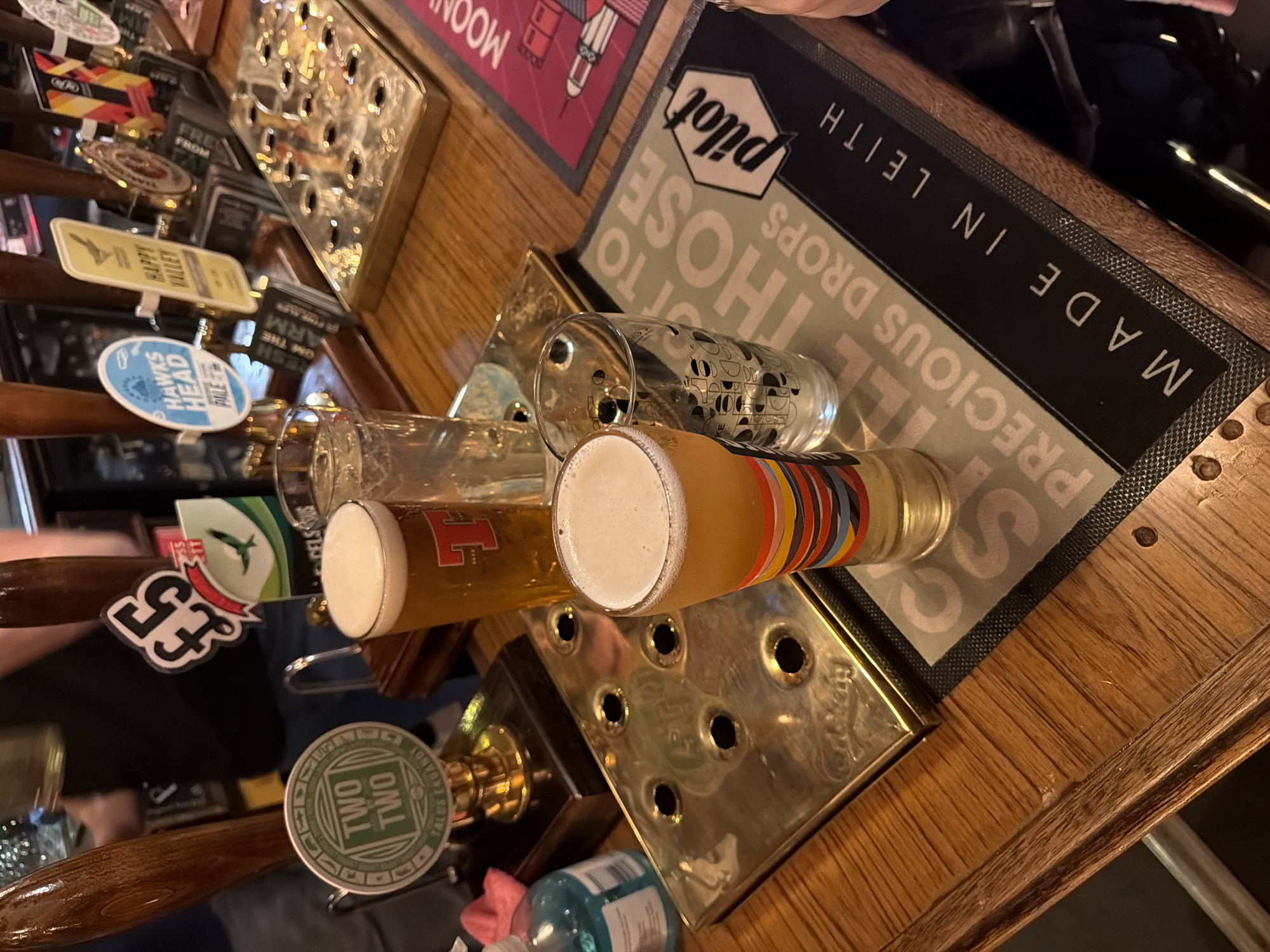 The scene depicts a polished wooden bar counter within a pub located in Leith, City of Edinburgh, United Kingdom. The counter displays a row of active beer taps, each with distinct pump clips, including "TWO TWO TALUS FRUMANOT," one marked "£5" with a green bird logo, "HAWKS HEAD PALE," and "HAPPY VALLEY." Four glasses are present: a Tennent's-branded pint glass filled with amber beer and a white head, a tall clear glass containing a lighter liquid, a smaller clear glass with a patterned design, and a pint glass with multicolored horizontal stripes on its base, also filled with amber beer and a white head. A rectangular brass drip tray, faintly embossed with "Carlsberg" and featuring multiple circular perforations, is centrally positioned. Underneath the striped pint glass is a black bar mat prominently featuring the "PILOT" logo, "PRECIOUS DROPS," and the phrase "MADE IN LEITH." A red mat with a partial "MOON" inscription is visible to the right. A light blue pump bottle, likely hand sanitizer, and a pink cloth are situated on the far left of the counter. A blurry figure in a dark t-shirt is visible behind the taps, and a human hand is partially visible on the far right of the frame.