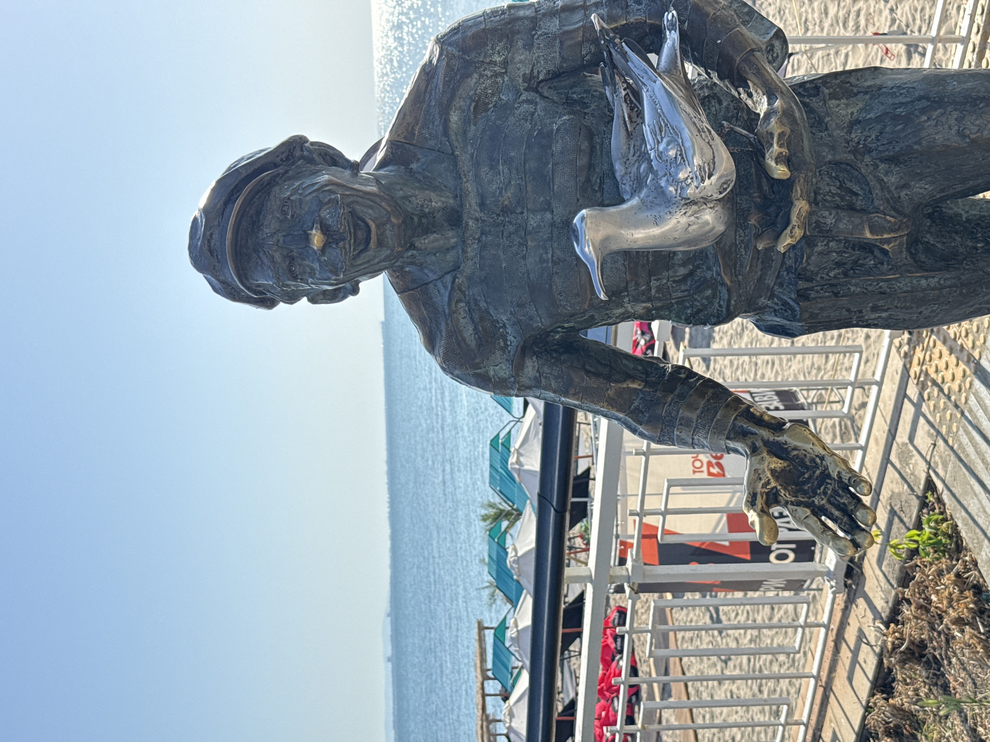 The image depicts a bronze statue of a fisherman holding a metal seagull, located on the Burgas Sea Garden in Burgas, Bulgaria. The statue faces the sea, with the sun glinting off the water and a clear blue sky above. In the background, beach chairs and umbrellas are visible, suggesting a recreational area. The statue is positioned on a paved walkway, with a white railing separating it from the sandy beach. The fisherman statue is rendered with detailed features, including a hat, a smiling face with a prominent nose, and a textured, striped garment. One hand of the statue is extended outwards, while the other holds the seagull. The overall scene is a snapshot of a sunny day at a seaside location.