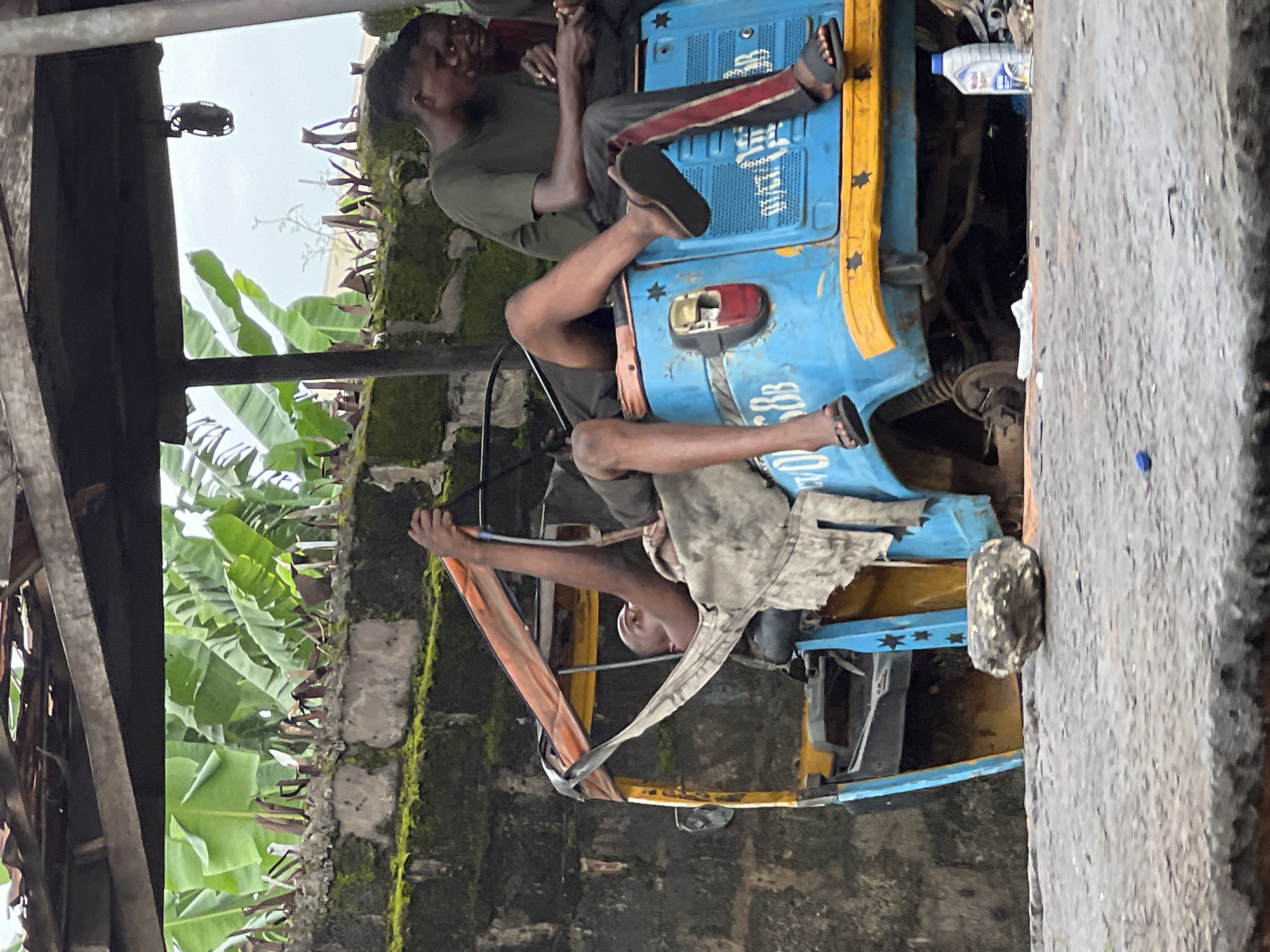 The image depicts two young males in Nkpor, Nigeria, appearing to be resting or working on a blue and yellow auto-rickshaw. One individual, dressed in a dark green t-shirt and dark shorts with red and black striped accents on the legs, is seated on the front of the auto-rickshaw. He is wearing dark sandals. His gaze is directed downwards, and his posture suggests a moment of rest or contemplation.
The second individual is positioned lower, seemingly within or beside the auto-rickshaw, and is wearing a light-colored, possibly grey, garment. His legs are visible, and he is also wearing dark sandals. He appears to be leaning against the vehicle or a part of its structure.
The auto-rickshaw itself is vibrantly colored, with a prominent blue body and a yellow trim. Stars are visible as decorative elements on the blue section. A red taillight is also apparent. In the background, lush green banana leaves are visible, suggesting a tropical or subtropical environment. Behind the leaves, a moss-covered brick wall and the sky are discernible. The overall scene suggests an outdoor setting, possibly a workshop or a roadside area, in what appears to be daylight, though the sky is overcast, implying a potentially humid or rainy weather condition. The individuals' expressions are not clearly discernible but do not convey overt distress. There is no visible text of significance, apart from some indistinct markings on the side of the auto-rickshaw.
