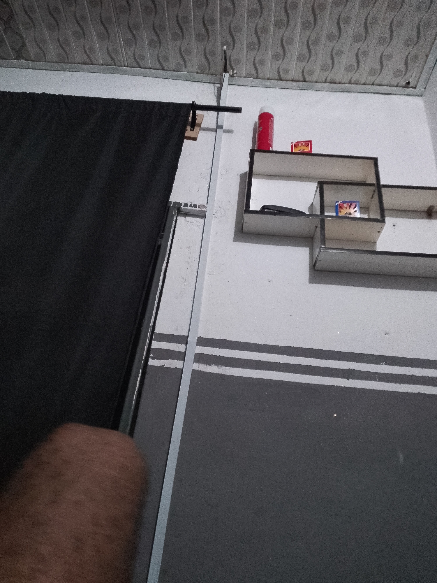 A low-angle, close-up shot shows a black curtain partially obscuring a wall. The wall features a white, L-shaped shelf unit holding a red aerosol can, a small red and yellow box, and a dark object, possibly a phone. Below the shelf, the wall is painted with two horizontal grey stripes. A ceiling with a patterned texture is visible at the top. A blurry, brown object, possibly an arm or leg, is in the lower left corner of the frame. The scene is indoors, suggesting a residential or commercial space within Jauro Yinu, Nigeria.