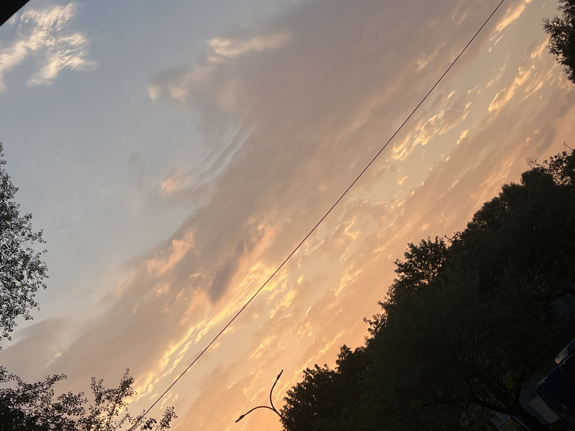 The image depicts a sunset with dramatic clouds in the sky. The sky transitions from a soft orange and yellow hue on the left to a pale blue on the right. wispy clouds, illuminated by the setting sun, are scattered throughout the sky. In the foreground, the dark silhouettes of trees line the bottom of the image, obscuring much of the ground. A street light with a curved pole is visible on the left side. A thin power line stretches diagonally across the frame from the top left to the bottom right. The image appears to be taken from a street-level perspective, looking upwards towards the sky, with hints of urban elements like buildings and vehicles barely visible in the lower left corner. The scene is likely set in a park or a tree-lined street within the city of Warsaw, Poland, where such natural light phenomena and urban infrastructure are common.