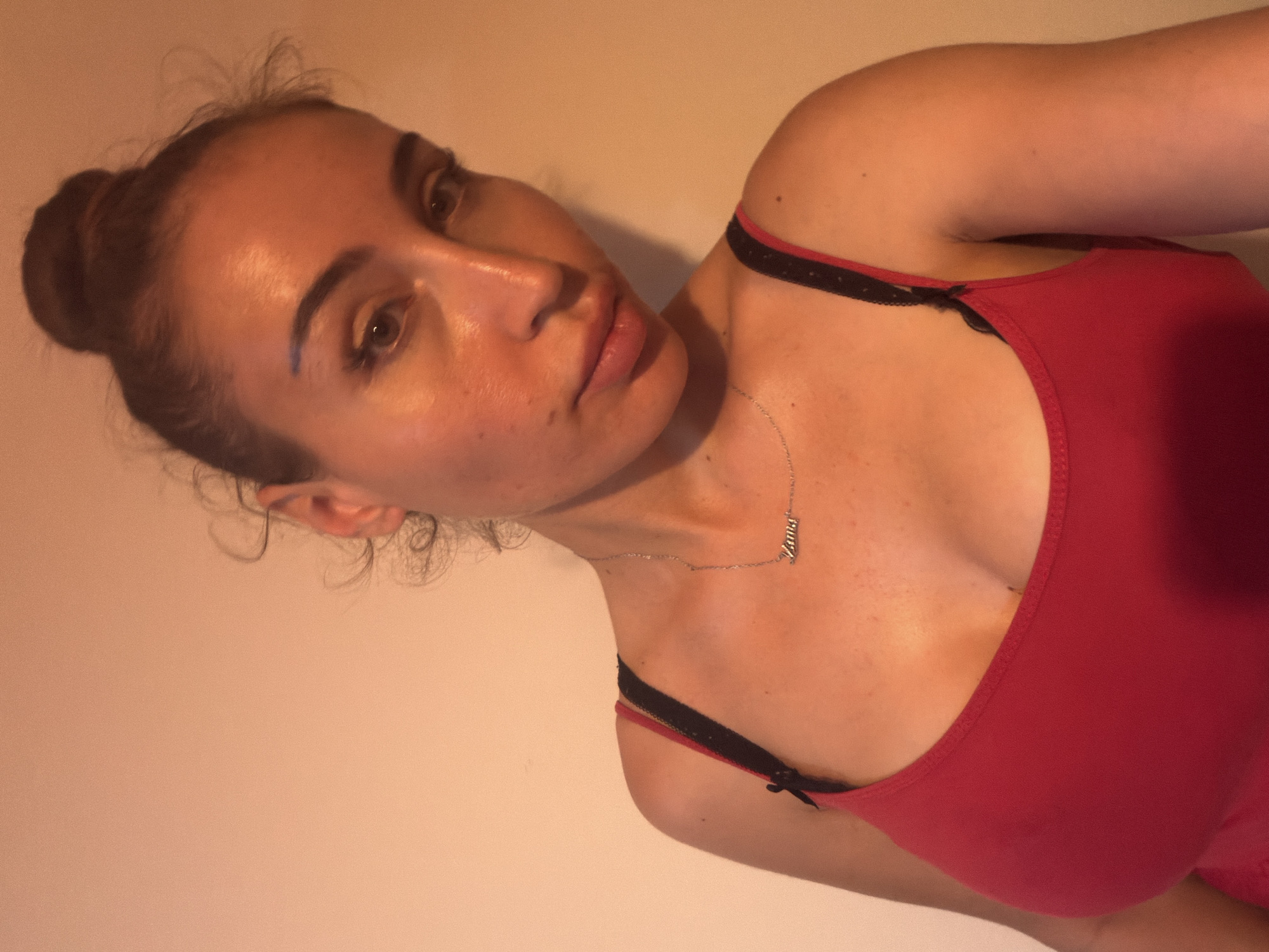 The media file is a close-up, angled portrait of a young woman with her hair pulled up in a bun. She is wearing a red tank top with black lace trim. A delicate silver necklace with a pendant is visible on her chest. The background is a plain, light-colored wall. The lighting suggests an indoor setting, possibly during the evening or under artificial light. The woman's gaze is directed towards the camera, and her expression appears neutral. The image focuses on her face, shoulder, and upper chest, highlighting her features and the jewelry. There is no visible text in the image. The location context is Tersefanou, Cyprus.