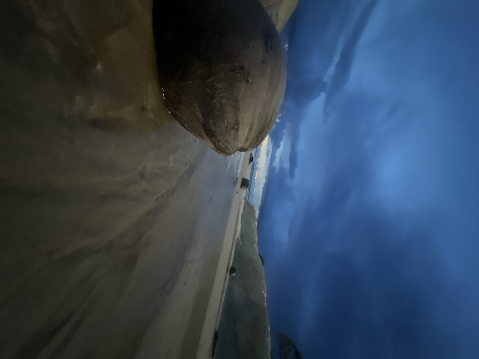 The image captures a close-up of a large, textured coconut resting on a wet, sandy surface. The coconut dominates the foreground, its rough, brown exterior partially submerged in shallow water. Behind the coconut, a sandy beach stretches out, meeting a dark, cloudy sky. The sky is a deep, moody blue, with swirling patterns suggesting impending rain or a storm. There are some faint lights visible on the horizon, possibly from a distant settlement or boats. The overall mood is dramatic and slightly ominous due to the dark sky and the way the light is hitting the scene. The time of day appears to be dusk or early evening, given the low light and the color of the sky. The location is identified as Choeng Thale, Thailand, which is known for its beaches and tropical climate. There is no visible text in the image.