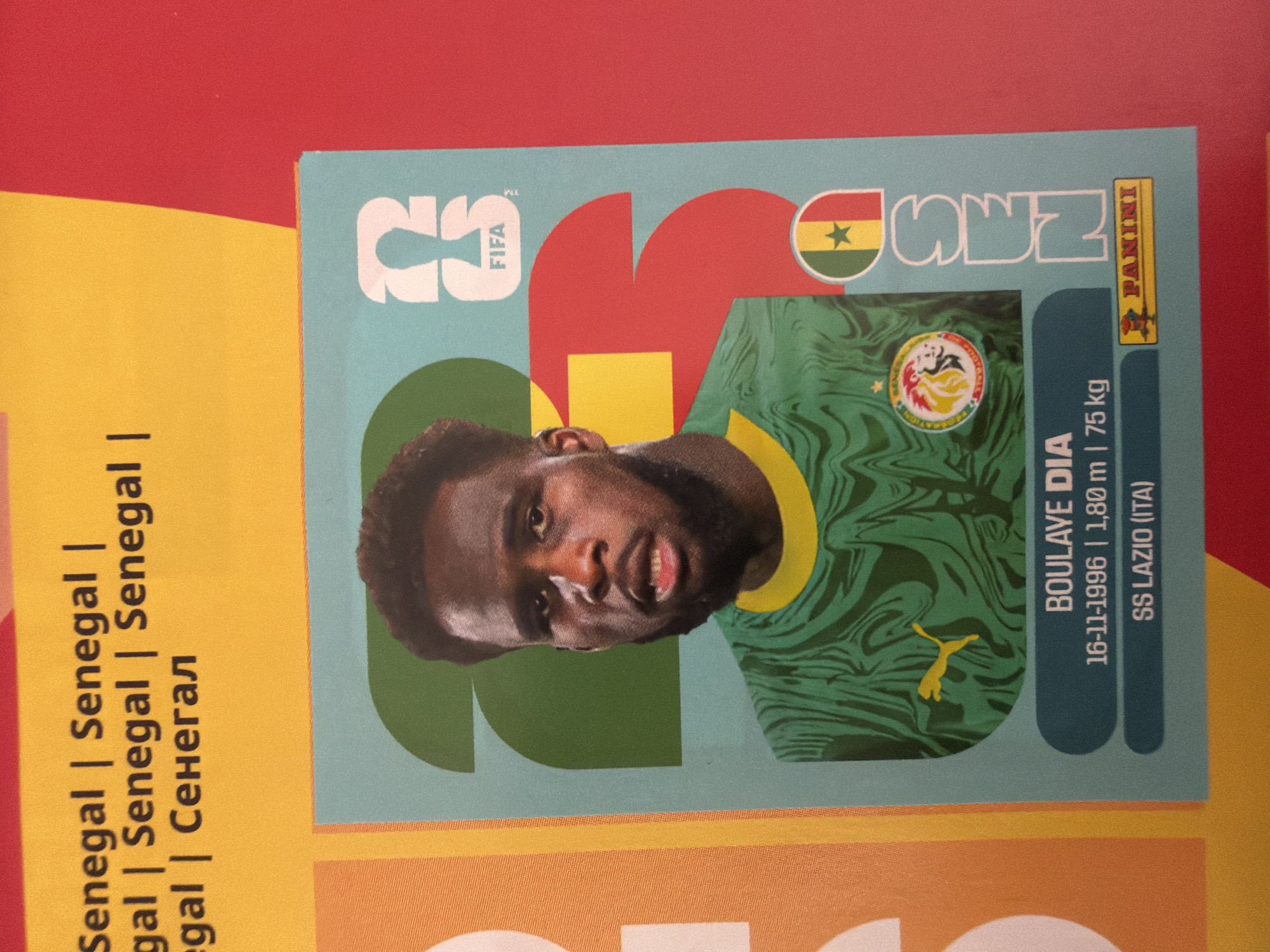 The image shows a close-up of a football sticker featuring player Boulaye Dia. He is wearing a green and yellow patterned jersey, the national colors of Senegal. The sticker is light blue with large, stylized white numbers that appear to be "2" and "0", possibly indicating the year 2022. The FIFA logo is visible near the top right. Below the player's image, his name "BOULAYE DIA" is printed, followed by his date of birth "16-11-1996", height "1,80 m", and weight "75 kg". His club, "SS LAZIO (ITA)", is also listed. A Panini logo is present at the bottom. The background of the sticker has geometric shapes in red and yellow. The sticker is placed on a surface that appears to be a red and yellow patterned booklet or album. Text in yellow at the top of the image reads "Senegal | Senegal | gal | Senegal | Senegal | egal | Сенегал". The location in the city of Zlatitrap, Bulgaria, is not evident from the image itself.
