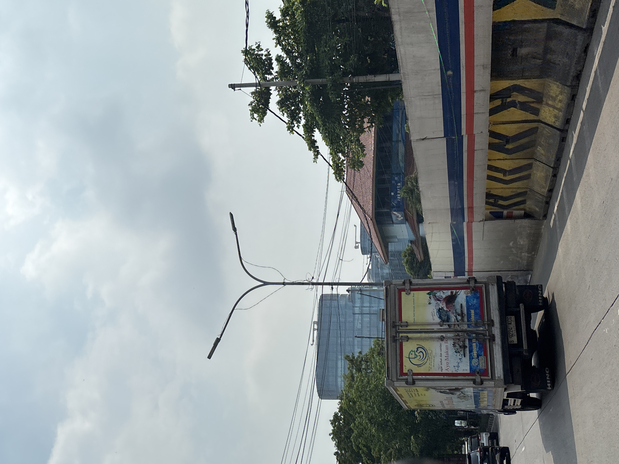 The image shows a street scene in North Jakarta, Indonesia. A truck with a white and yellow cargo container is on the road. The cargo container has several advertisements on it. In the background, there's a modern glass building, trees, a street light, and an elevated concrete structure painted with blue, white, and red stripes and yellow and black chevrons. The sky is cloudy. The image appears to be taken from a low angle.