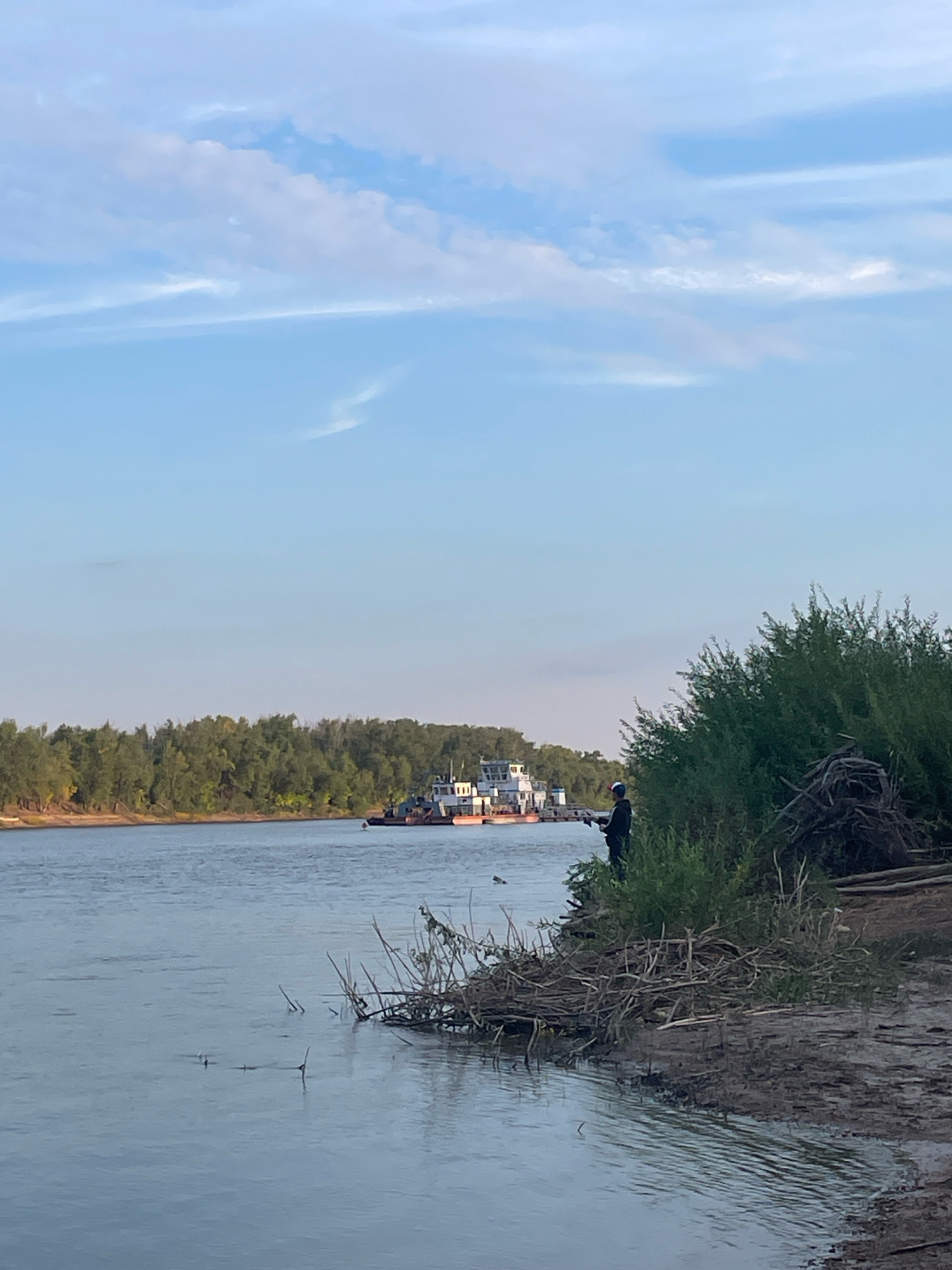 The image depicts a wide, calm river with a barge and its tugboat in the middle ground, moving from left to right. On the right bank, a person stands near the water's edge, facing the river. The bank is covered with vegetation and debris. The left bank is lined with a dense forest. The sky is partly cloudy with a clear blue expanse. The scene appears to be a natural landscape with human activity, possibly in or near the city of Oral, Kazakhstan, given the river's prominence.