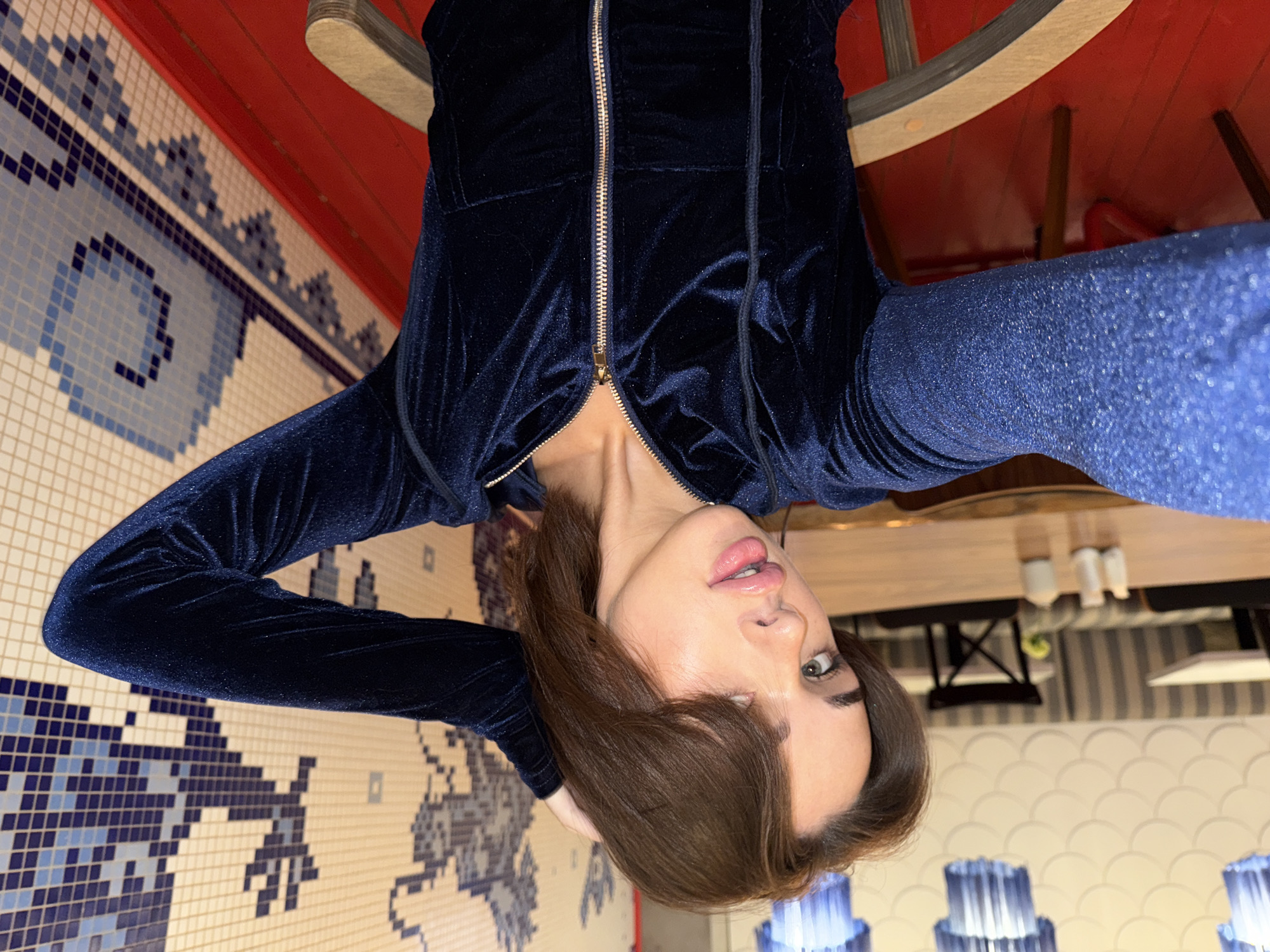 This image is a selfie of a person, likely female, with medium-brown hair, green eyes, and glossy lips, looking directly towards the camera with a neutral expression. She is dressed in a dark blue velvet or velour hooded jacket with a partially unzipped gold zipper, revealing a hint of her upper chest. Her right arm, covered in a glittery blue long-sleeved garment, is extended, while her left hand is positioned near her head.
The background on her left is a wall adorned with an intricate blue and white mosaic tile pattern, showcasing geometric designs and stylized figures. On her right, the indoor setting, likely a restaurant or cafe in Perm, Russia, includes a red ceiling or partition, and parts of wooden tables and chairs. Further in the background, a light-colored wall displays a subtle, textured pattern. Several distinctive light fixtures with blue, ribbed shades are visible, casting a soft blue glow. The lighting suggests an evening or night setting, or a well-lit interior space. The image is originally presented rotated 90 degrees counter-clockwise. No text is visible.