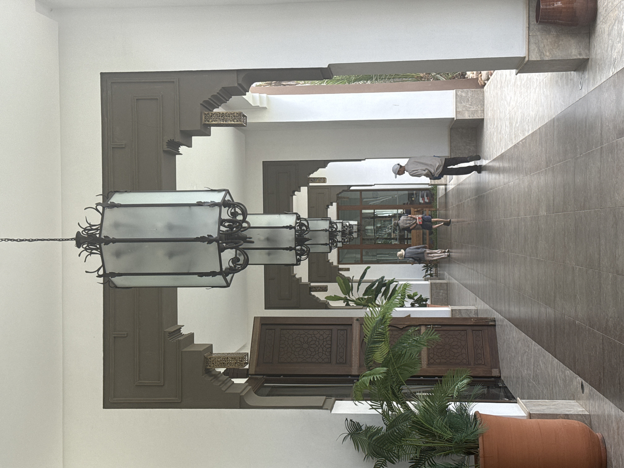 The image shows an interior hallway, likely in Agadir, Morocco, based on architectural style. The hallway features decorative archways and doors, alternating on either side. An ornate, multileveled chandelier hangs from the ceiling. A potted plant is near the bottom right corner. Several people are walking down the hallway in the distance. The floor is tiled in neutral tones. The walls are white, contrasting with the darker archways.