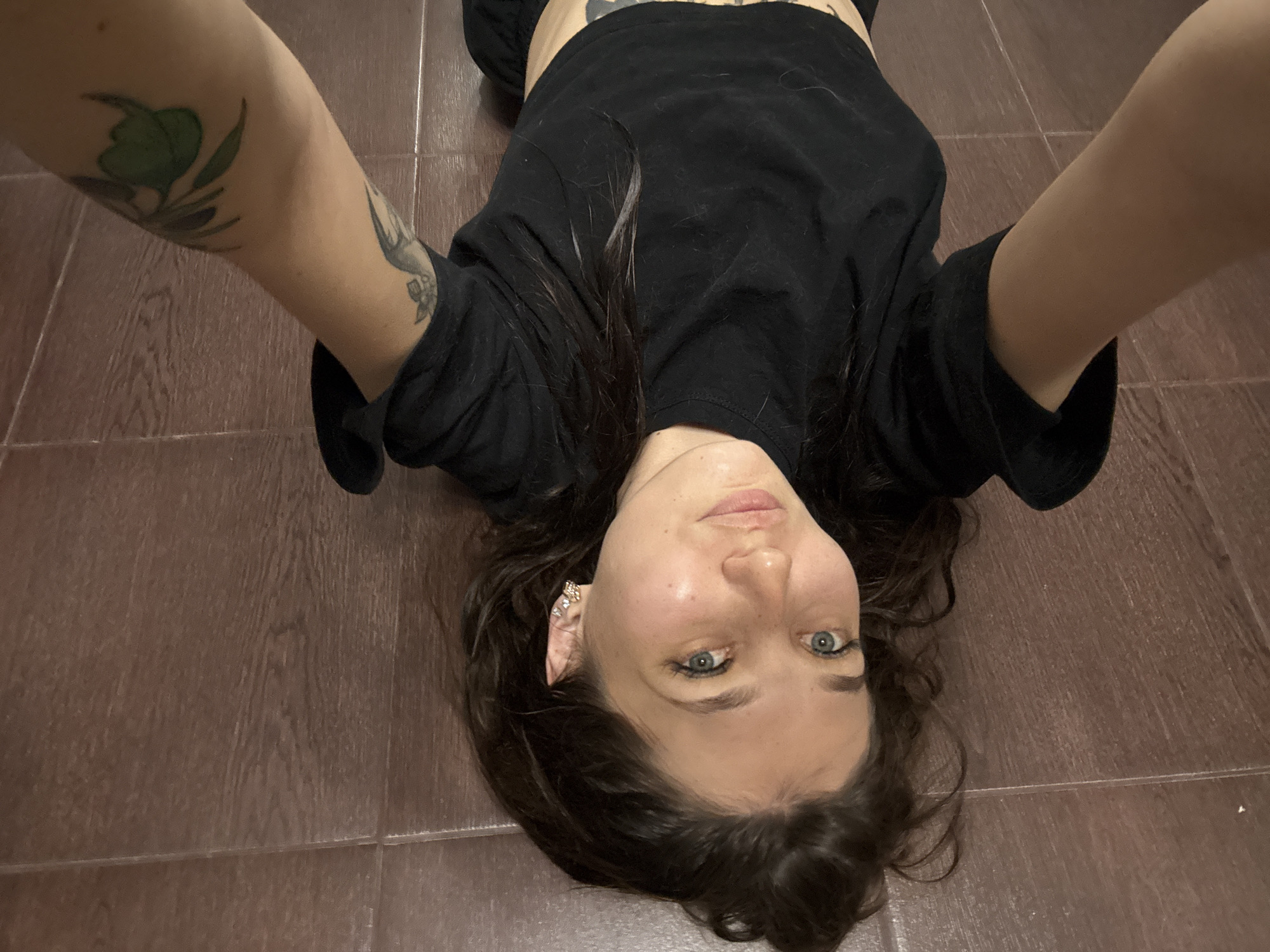 A young woman is lying on her back on a wooden floor, her head towards the camera. She is wearing a black t-shirt and dark pants. Her long, dark hair is spread around her head. She has a tattoo of a green leaf and stem on her left arm. She is wearing a sparkly earring in her left ear. Her eyes are blue and she is looking at the camera with a neutral expression. The lighting appears to be indoor artificial light, and no specific time of day or weather conditions can be determined. The setting is likely a home or indoor space with a dark brown tiled floor. No visible text is present in the image.