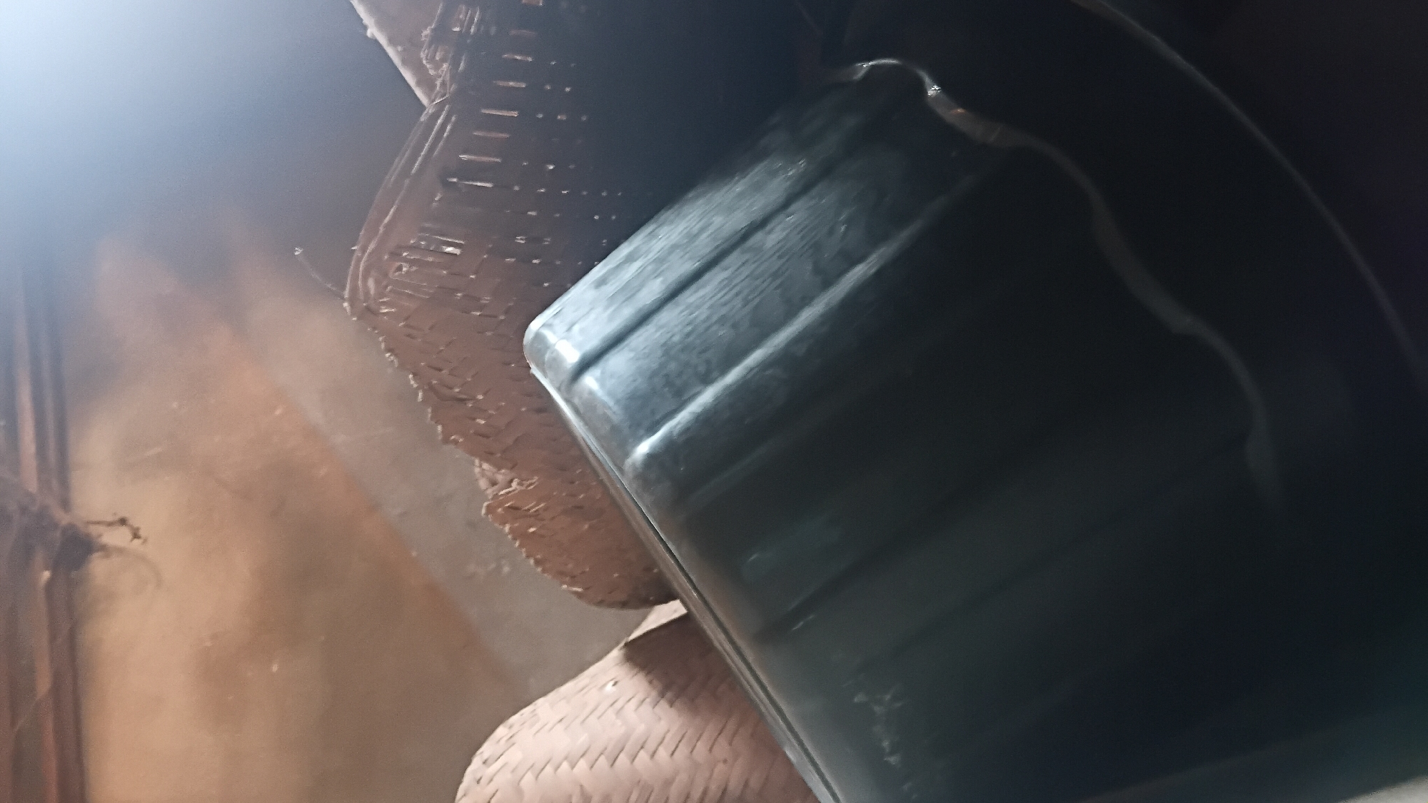 The image shows a close-up view of several wicker baskets stacked together, with a dark, glossy plastic container in the foreground. The baskets appear to be made of natural fibers and have a woven texture. The plastic container is large and rectangular with ribbed sides. The background is dimly lit and appears to be an interior space, possibly a storage area, with a dusty or textured wall. The lighting suggests it may be nighttime or an indoor space with artificial light, casting shadows and highlighting the textures of the objects. The scene is devoid of any human presence. The location of this scene in Prabumulih, Indonesia cannot be determined from the visual information provided in the image.