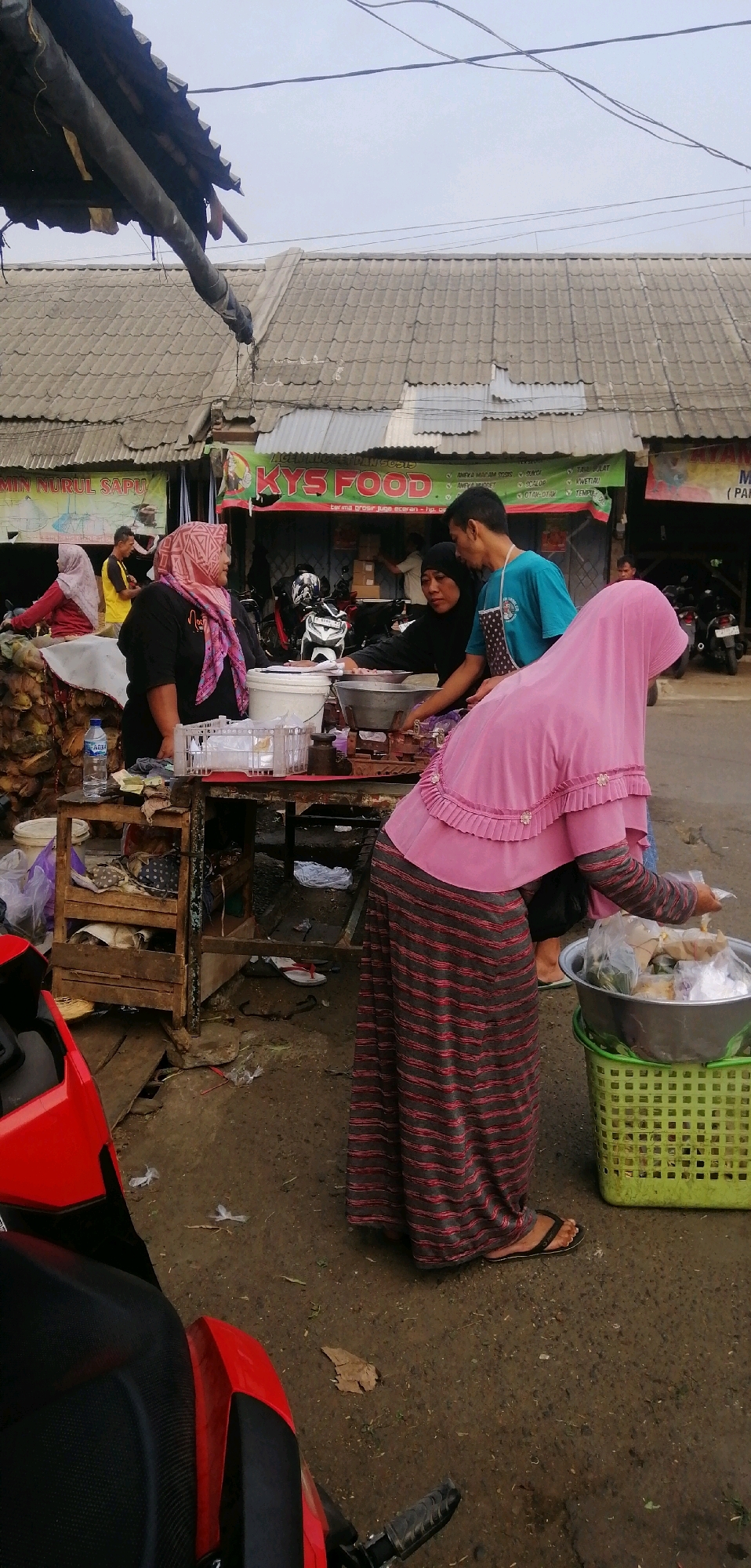 The image depicts a bustling outdoor market scene in Ajibarang, Indonesia, likely in the morning or late afternoon given the soft, diffused lighting. A vendor, a woman wearing a pink hijab and a striped dress, is actively serving customers at a wooden stall. She is interacting with a man in a teal shirt and a black headscarf, who appears to be a fellow vendor or customer. Another woman, also in a hijab and black attire, is visible behind them, tending to the stall. In the foreground, a portion of a red motorcycle is visible, suggesting the presence of transportation. The background reveals a row of shops with tiled roofs and awnings, one of which displays a prominent sign reading "KYS FOOD". The ground is a mixture of dirt and some debris, indicating an unpaved market area. The overall atmosphere is one of commerce and daily life in a local Indonesian community.