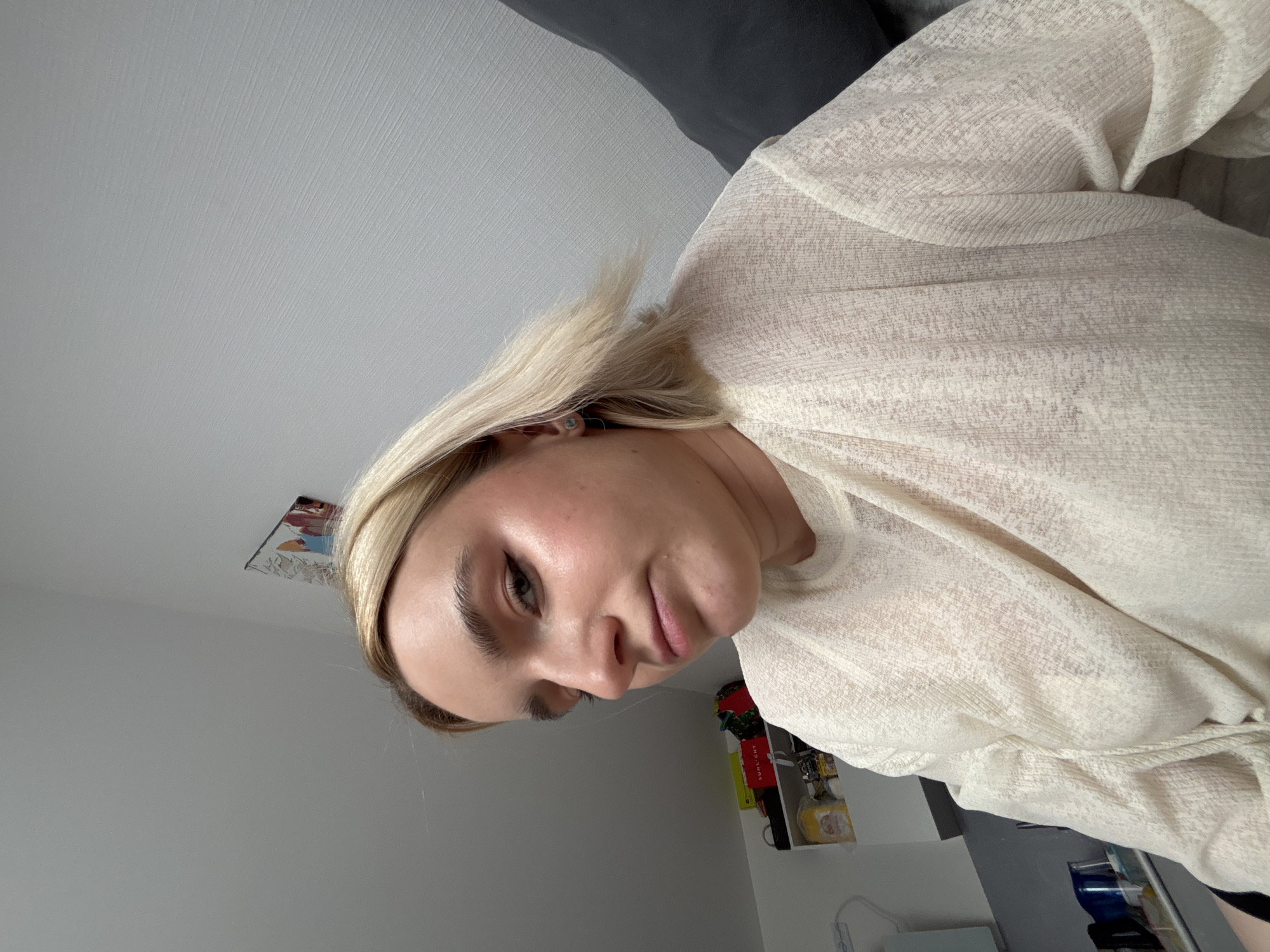 A woman with blonde hair is facing the camera. She is wearing a light-colored shirt. The background includes a gray wall and a shelf with various items. The location is Vsevolozhsk, Russia.