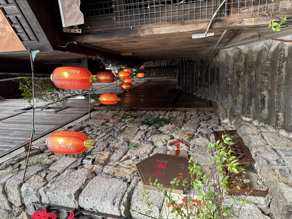 The image depicts a narrow, ascending stone staircase alleyway in Gongtan, China, flanked by a rough-hewn stone wall on the left and a dark wooden building on the right. A series of illuminated red Chinese lanterns of various sizes are suspended overhead, strung between the buildings, casting a warm glow. The stone steps appear wet and uneven.
On the left stone wall, a rectangular brown sign with white Chinese characters, possibly reading "鱼苑" and "古镇·古味" (Ancient Town, Ancient Flavor), is mounted. A red arrow is painted on the stone below this sign. Below the sign, a dark brown wooden planter box with a lattice design holds green leafy plants. Further left and partially cropped, a larger red sign with white characters is visible. A small grey electrical box is attached to the stone wall near the top left lantern.
The wooden building on the right features dark vertical planks and a window with a metal grid near the bottom right. Further into the alley, a dark wooden doorway is visible. Small green foliage grows from crevices in the stone wall and along the wooden structure. No individuals or animals are present.