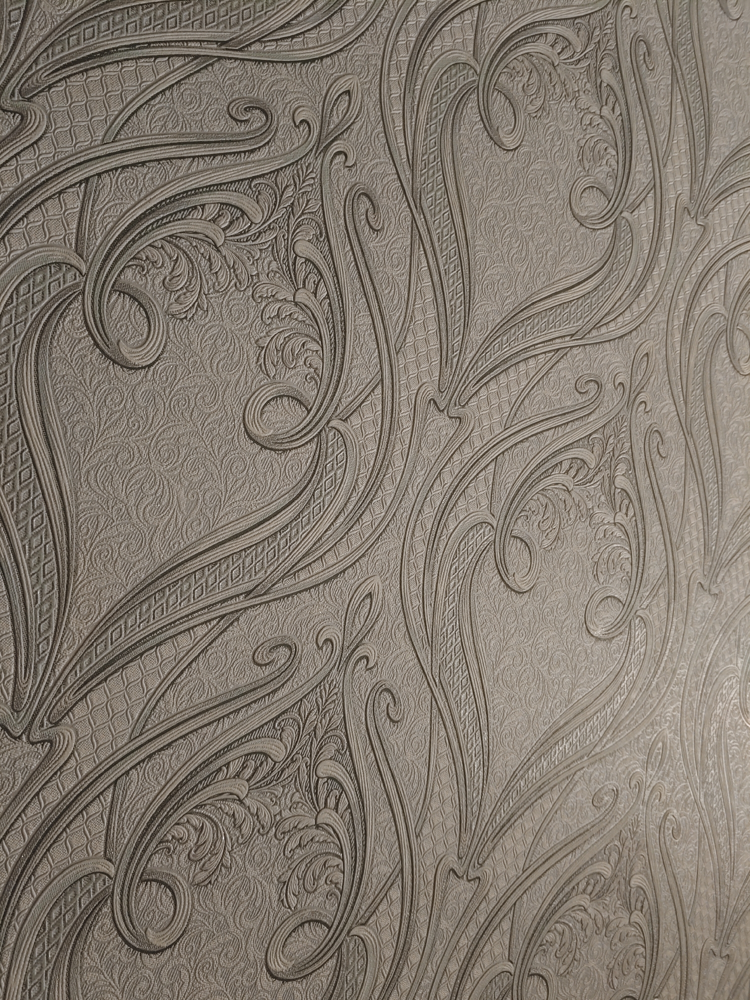 The image shows a close-up, diagonal view of embossed wallpaper with an ornate, scrolling floral pattern. The pattern is rendered in a monochromatic, muted gray or taupe color, giving it a subtle and sophisticated appearance. The texture of the wallpaper is visible, with raised lines and delicate detailing creating a sense of depth and dimension. The background has a finely textured, almost damask-like pattern, over which the bolder, raised design is superimposed. The lighting emphasizes the relief of the pattern, casting soft shadows that highlight the contours of the scrolls and floral elements. There are no people or discernible objects within the scene, making it a purely textural and decorative study. The location of Курашим, Russia, is not directly indicated by any visual cues in the image itself.