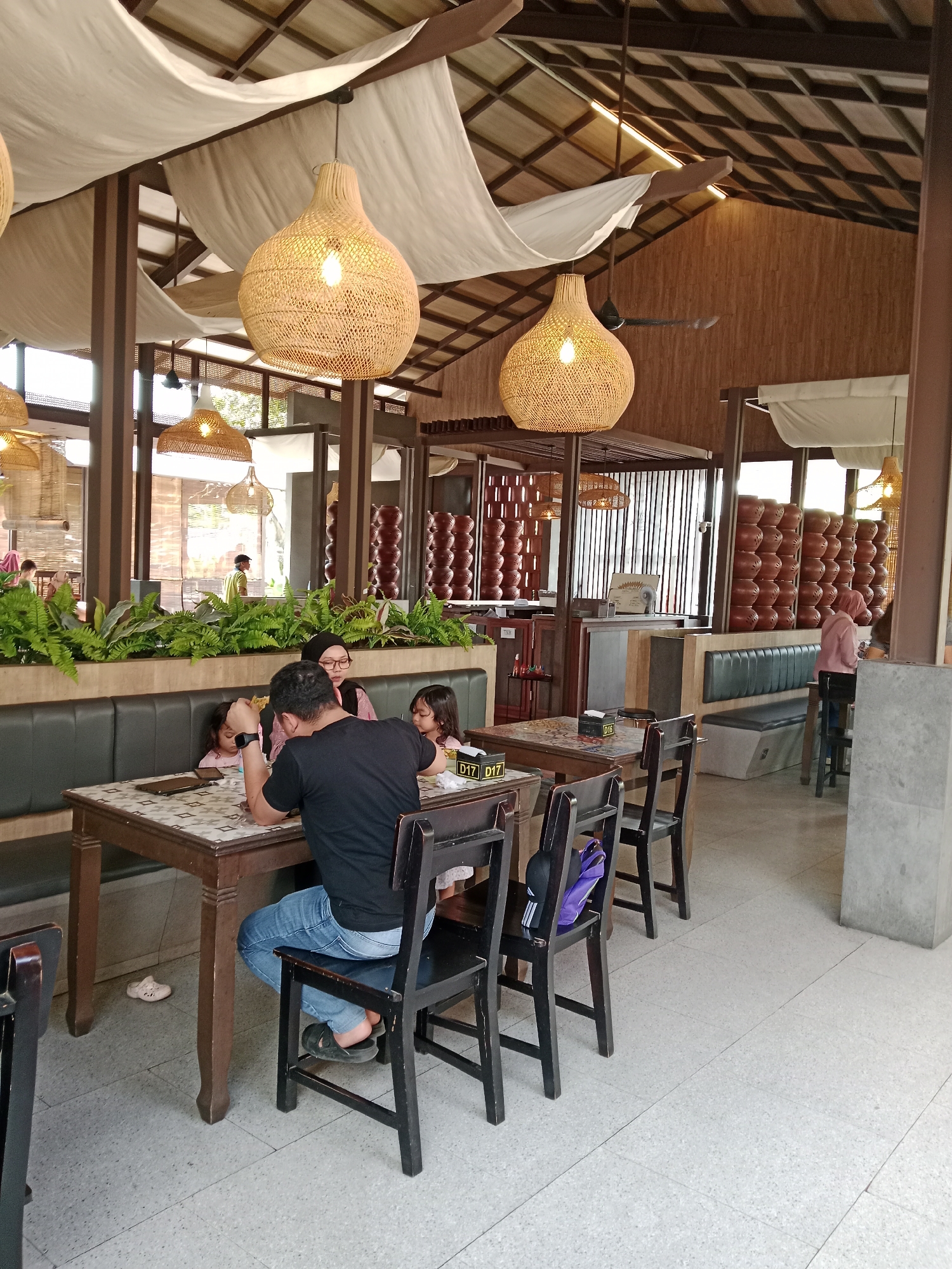 The image depicts a family of four seated at a wooden table in a restaurant with a rustic, natural theme. A father wearing a black t-shirt and blue jeans is seated facing away from the camera, looking at the table. His wife, wearing glasses and a pink headscarf, sits to his left, facing the camera. Two young girls, presumably their daughters, are seated on the other side of the table. One girl, wearing a pink top, is looking at the table, while the other, also in pink, is partially visible and appears to be eating. The table is set with a patterned tablecloth, and on it are plates, cutlery, and napkins.
The restaurant interior features a high ceiling with exposed wooden beams and bamboo accents, draped with white fabric panels. Several large, woven pendant lights hang from the ceiling, casting a warm glow. Lush green plants are arranged along a booth seating area behind the family. In the background, rows of brown ceramic pots are stacked on shelves, and a person in a pink top is visible walking in the distance. The flooring is a speckled terrazzo tile. The restaurant appears to be open-air or have large windows, as indicated by the natural light entering the space. The overall ambiance suggests a relaxed dining environment, possibly in Kecamatan Pati, Indonesia, given the architectural style and natural elements.