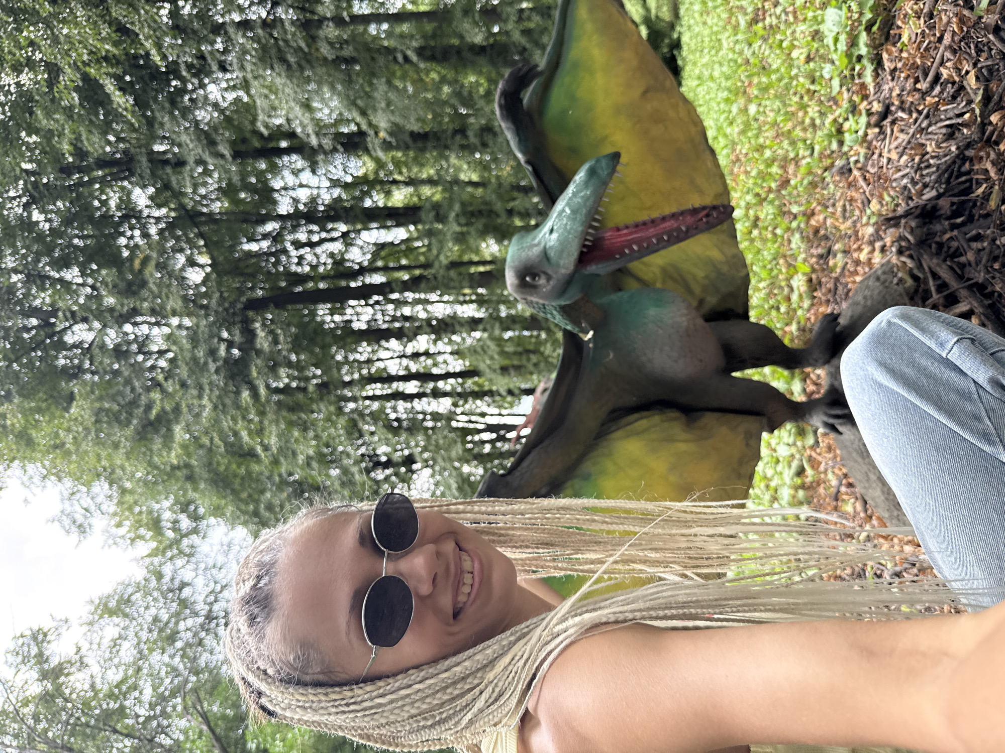 In Rusanovo, Russia, a young woman with blonde braided hair and sunglasses smiles at the camera in a wooded area. To her right, a realistic dinosaur model with green and yellow wings and its mouth open is positioned. The woman is wearing a light-colored top and blue jeans. The ground is covered with leaves and twigs, and the background features a dense forest.