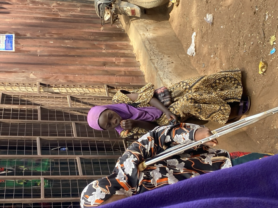 The image displays a street scene in Mokwa, Nigeria, under daylight conditions. In the left foreground, the upper back and right arm of a person, likely female, are visible, clad in a black head covering and garment with white and orange floral patterns. This individual holds a silver crutch with their right hand. To their right, in the midground, a young female wearing a purple hijab and a long, yellow and black patterned dress is seated on a low concrete ledge. She holds her hands near her mouth, seemingly consuming a small, orange object. A small, striped shoulder bag hangs from her left shoulder, and she wears purple open-toe sandals. The ground beneath the ledge is dusty, reddish-brown earth, scattered with pieces of white plastic, a blue wrapper, and other debris. The background on the left features a dark, gridded metal structure, while the right background consists of a weathered, reddish-brown corrugated metal wall with a white paper or poster affixed to it. A portion of a motorcycle's rear wheel and fender is visible on the far right.
