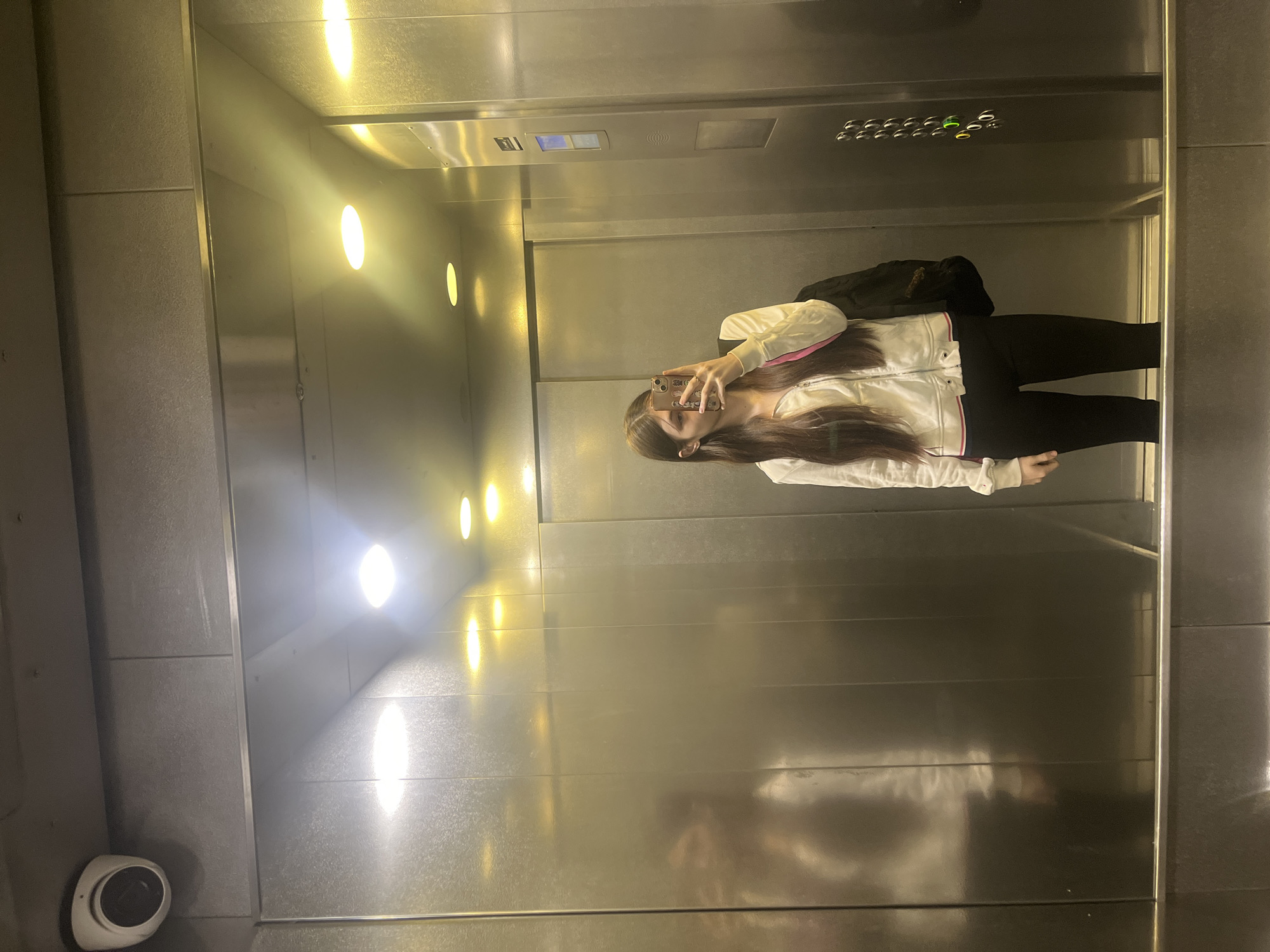 An adolescent female stands in an elevator, holding a smartphone to capture a selfie. She's clad in black leggings and a white jacket with pink accents, also sporting a black backpack. The elevator's interior is illuminated by a row of overhead lights, casting a reflective sheen on the metallic walls. At the lower left of the image, a security camera is mounted. The location is potentially within Saint Petersburg, Russia, but requires further evidence for definitive confirmation.
