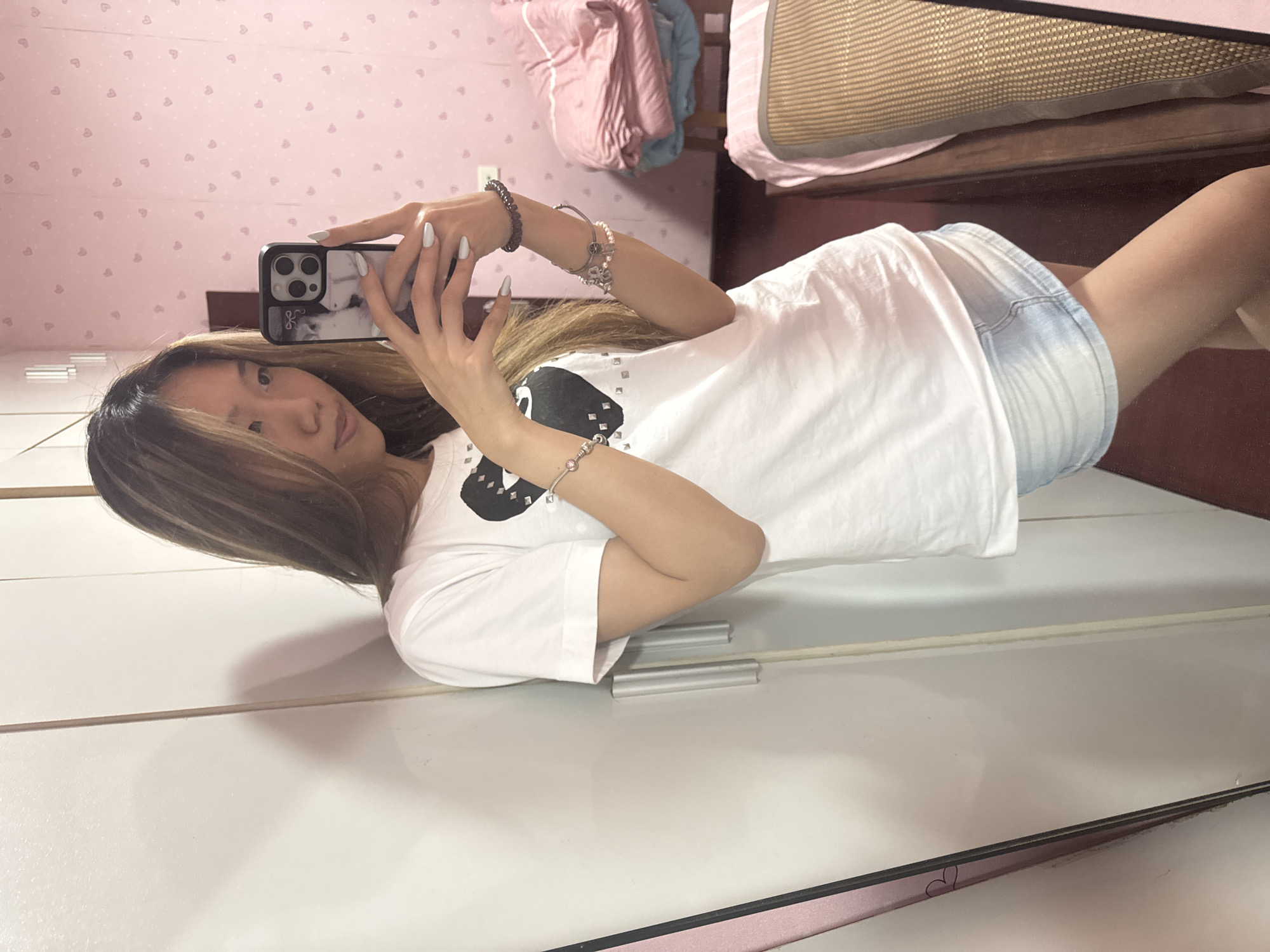 A young woman is taking a selfie using her mobile phone with a light-colored case in what appears to be a bedroom in Shapingba District, China. She is wearing a white t-shirt with a black design and light blue denim shorts. She has bracelets on her wrists and is standing in front of a white wardrobe. In the background is a bed with pink bedding. The wall is decorated with a small heart pattern.