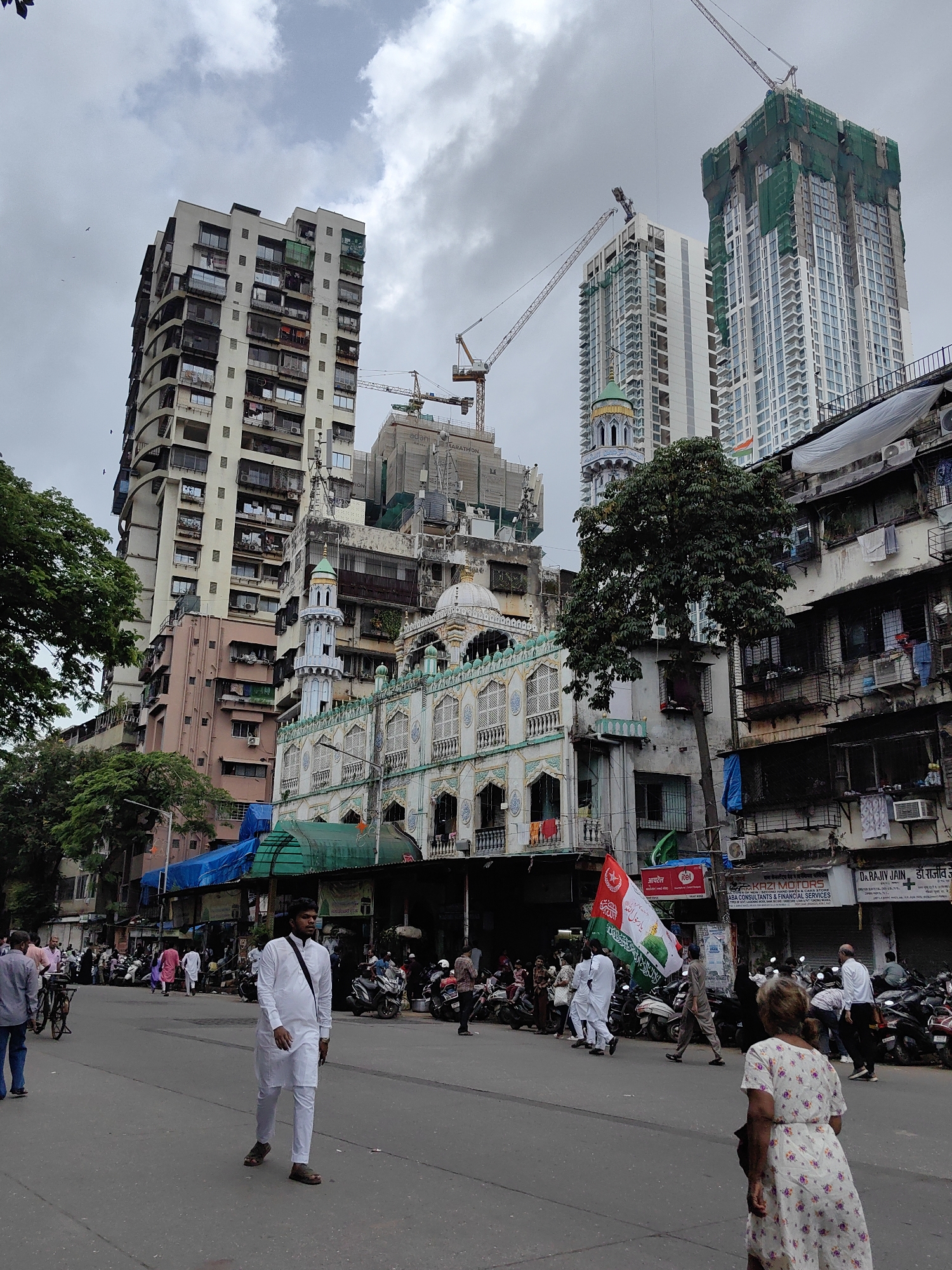 The image depicts a street scene in Mumbai, India, with a prominent mosque in the foreground and several high-rise buildings, some under construction, in the background. The sky is overcast, suggesting cloudy weather.
In the foreground, a man in a white kurta pajama walks towards the right of the frame. To his right, a woman in a floral dress is partially visible, facing away from the camera. Behind them, a street is lined with parked motorcycles and scooters. A large green flag with Arabic script and what appears to be the Indian flag are being carried by people further down the street, in front of the mosque.
The mosque itself is an ornate structure with a green dome, white minarets, and decorative architectural elements. Adjacent to the mosque, older, multi-story buildings with balconies and window air conditioning units are visible, showcasing a mix of architectural styles and the dense urban environment. Further back, towering skyscrapers, some shrouded in green construction netting, dominate the skyline, indicating ongoing development in the city. Trees are interspersed throughout the scene, providing some greenery amidst the concrete and steel. The overall impression is a bustling urban environment with a blend of religious architecture, residential buildings, and modern construction.