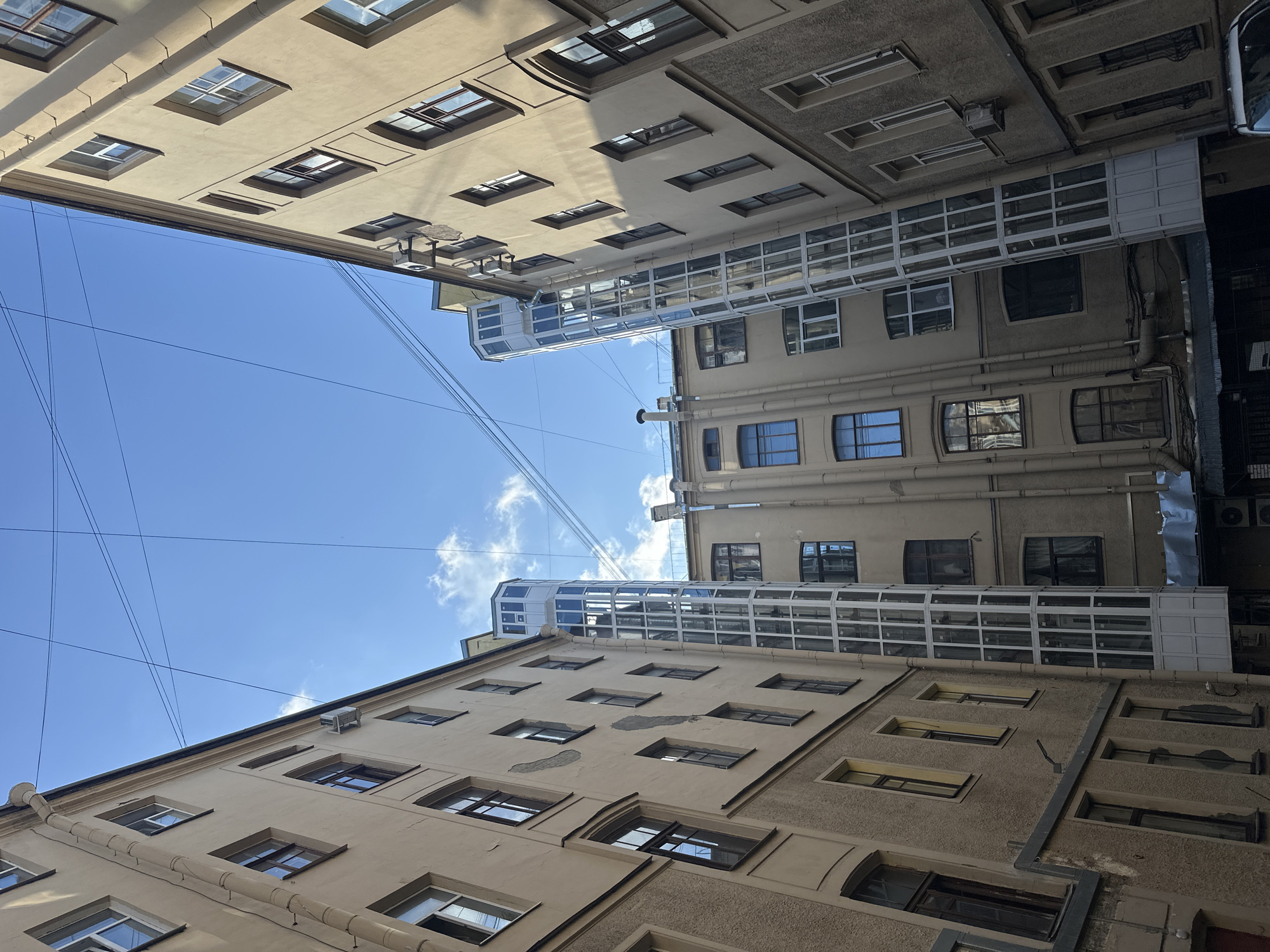This image captures a narrow courtyard in Saint Petersburg, Russia, with tall, aged buildings on either side. The architecture suggests a historical district, likely featuring Soviet-era or earlier residential buildings. The buildings are constructed from light-colored stucco and have numerous windows of varying sizes and designs, some with dark frames. Two modern glass elevators are installed on the exterior walls of the buildings, reaching upwards towards the sky. Wires crisscross the sky, typical of urban environments. The sky is blue with scattered clouds, indicating it is daytime. The scene is devoid of people, vehicles, or any immediate signs of activity, presenting a quiet, architectural study of an urban courtyard. The perspective is looking upwards, emphasizing the height of the buildings and the enclosed nature of the space. The overall impression is one of a utilitarian urban environment with elements of both old and new architecture.