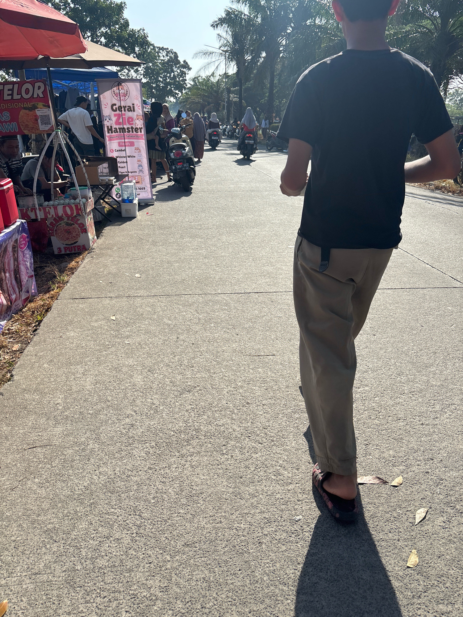 The image depicts a street scene in Telagamurni, Indonesia. In the foreground, a person wearing a black t-shirt and tan pants walks away from the camera, casting a shadow on the concrete pavement. To the left, several market stalls are set up along the road. One stall has a red umbrella and displays signs for "TELOR" and "AS LAWI". Another stall features a banner for "Gerai Zie Hamster" with cartoon images of hamsters and text listing hamster breeds. People are seen browsing at the stalls, and a few motorcycles are parked or passing by. Palm trees line the background, and the sky is clear and bright. The overall atmosphere suggests a bustling outdoor market on a sunny day.