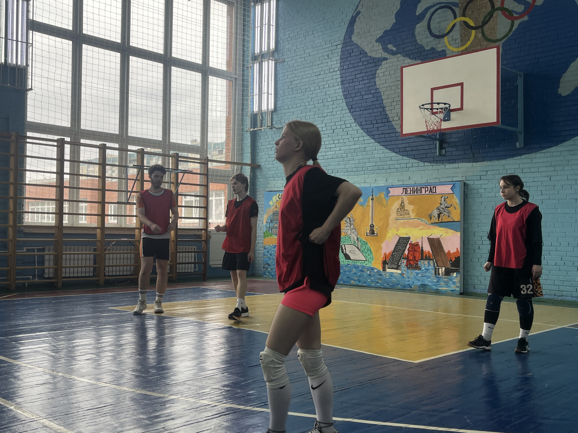 In a gymnasium setting, four individuals are present, all clad in attire suggesting involvement in sports activity, possibly volleyball. The three individuals in the background are positioned at the front of the room, while a fourth stands in the foreground. A tall window is behind the individuals in the background. Visible on the wall are depictions suggesting the location of Saint Petersburg, Russia, notably including an image of the Palace Bridge. Above the artwork is a mural that depicts a world map and the Olympic rings. The room also contains a basketball hoop mounted on the wall.