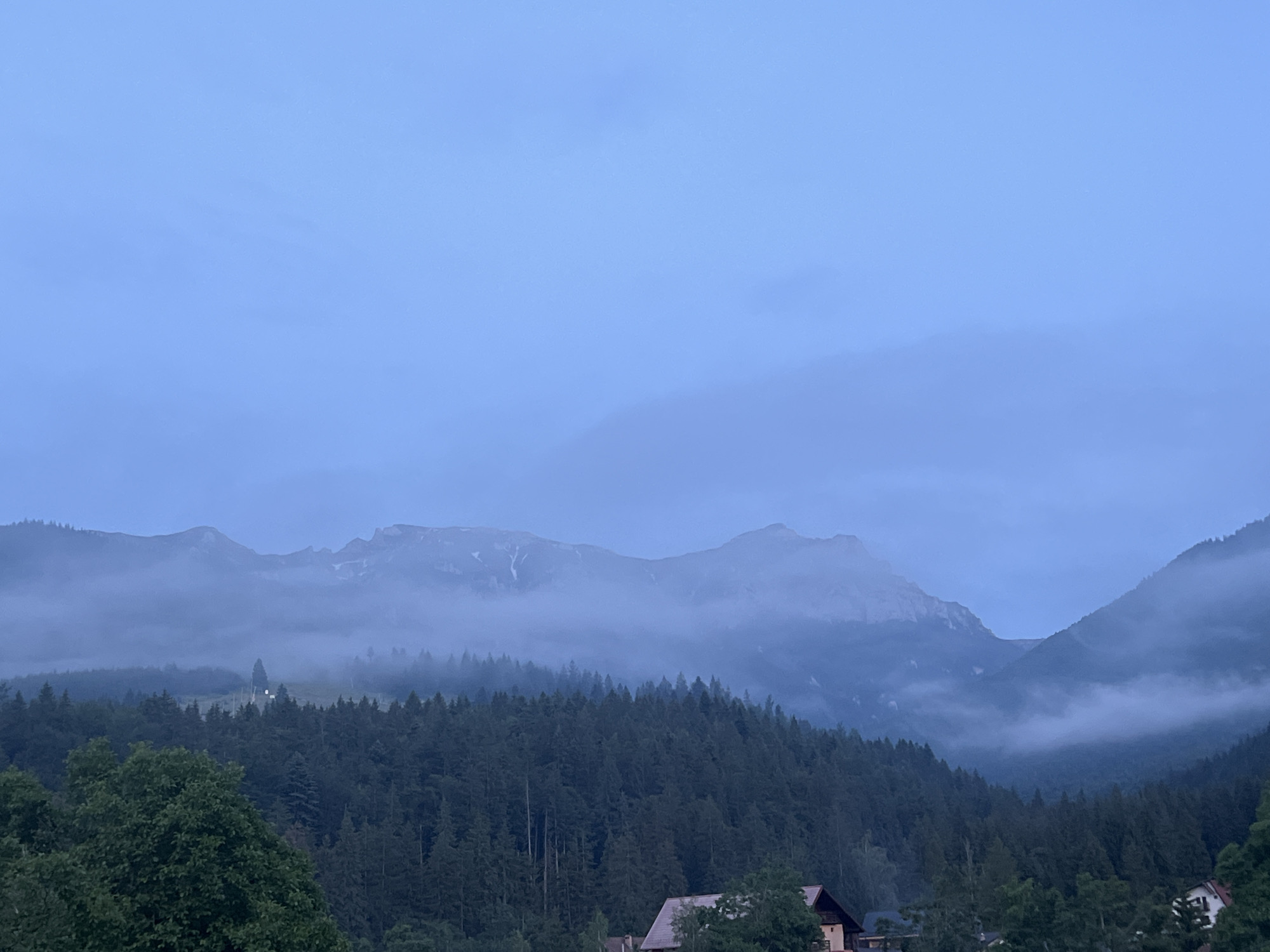 This is a wide-angle, horizontal, medium-shot photograph capturing a mountainous landscape at dusk or dawn. The sky is a pale, overcast blue, with a hint of haze or light fog. In the middle ground, a dense forest of dark green coniferous trees covers a hillside. Above the treeline, the rugged peaks of mountains rise, partially obscured by swirling mist and clouds. Some snow patches are visible on the higher mountain slopes. In the foreground, the tops of a few deciduous trees with green leaves are visible on the left side. Towards the bottom right, the peaked roof of a building, appearing to be a house with a light-colored wall, is peeking through the trees. The overall mood is serene and somewhat mysterious, with muted colors and diffused light. The scene is devoid of any people. The location is listed as Unknown, Unknown.