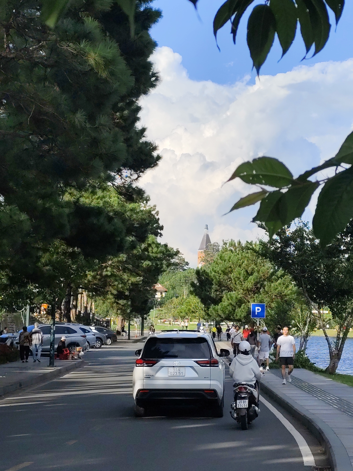 This image depicts a street scene in Xuân Hương Lake area of Da Lat, Vietnam. The foreground shows the rear of a white SUV and a person on a scooter. The street is lined with parked cars and a sidewalk. A blue parking sign with a white "P" is visible. People are walking on the sidewalk and in the background, there are trees and a lake. A building with a distinctive tower can be seen in the distance, behind the trees. The sky is partly cloudy.