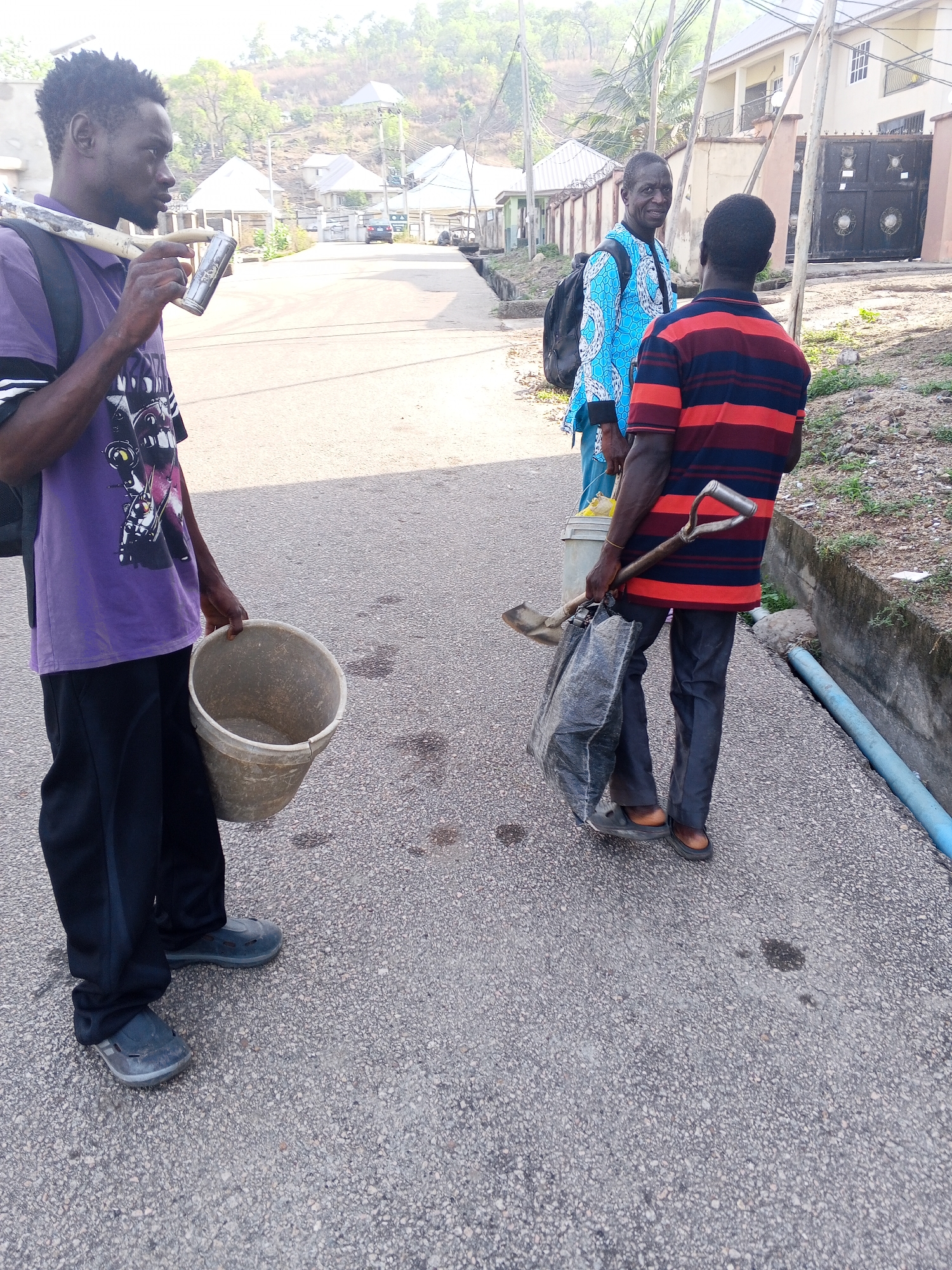 Three men are standing on a paved road in Lokoja, Nigeria. The man on the left is wearing a purple t-shirt, black pants, and grey shoes, carrying a bucket in his right hand and a long pole over his left shoulder. He has a black backpack on. The man in the middle is wearing a light blue patterned shirt and carrying a bucket. The man on the right is wearing a red and dark blue striped shirt, black pants, and brown shoes. He is holding a shovel and a black garbage bag. They appear to be sanitation workers. In the background are houses and green hills.