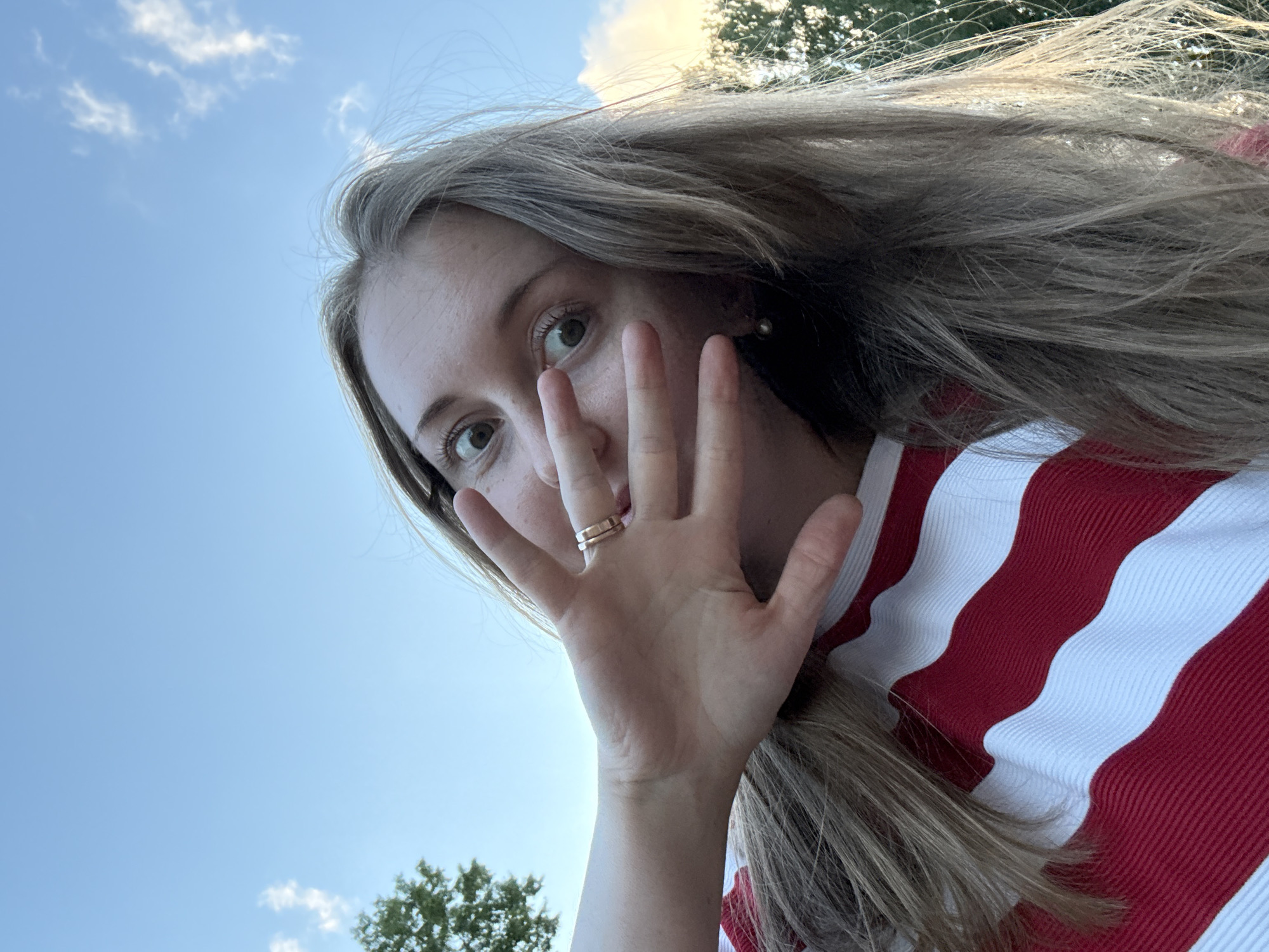 A young woman with long blonde hair and wide, surprised eyes covers part of her face with her left hand. She is wearing a red and white striped shirt and two rings on her left ring finger. The background is a clear blue sky with a few wispy clouds and the tops of trees visible. The image appears to be a selfie taken outdoors.