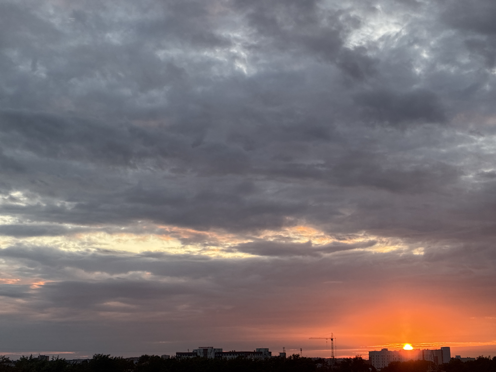 A wide shot captures a vibrant sunset over a city skyline. The sky is a dramatic canvas of dark, heavy clouds in the upper half, transitioning to lighter grays and then to a fiery orange and yellow gradient towards the horizon. The sun itself is a bright, glowing orb, partially obscured by the clouds, casting a warm light. Below the sunset, a silhouetted treeline marks the edge of the city. Buildings, including a prominent tower crane, are visible against the colorful sky, with the tops of several structures silhouetted against the orange glow. The overall scene evokes a sense of transition, with the end of the day marked by a spectacular display of natural light.