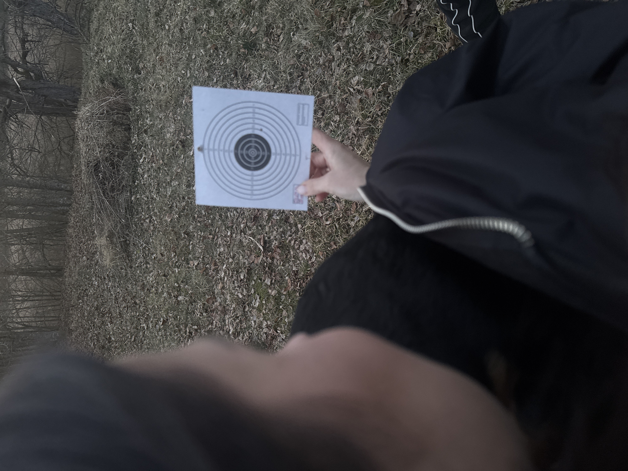 The image depicts a person holding a target in a rural area with patchy, brown vegetation, likely in the fall or early spring. The person is wearing a black jacket with white trim and black pants. The target is a standard circular shooting target with concentric rings. The foreground contains a blurry view of the person's face. The image was taken in Парэчча, Belarus.