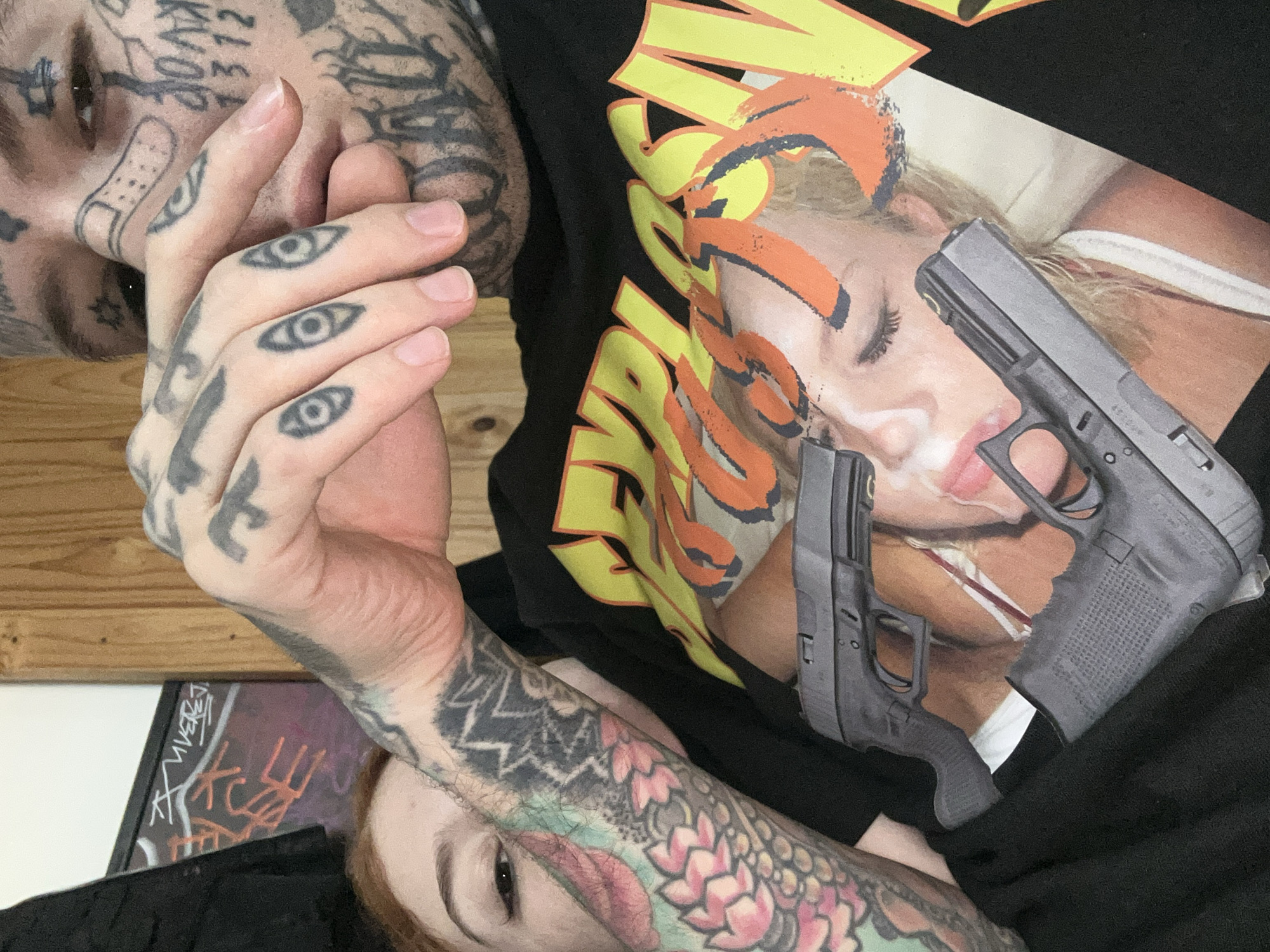 The image is a close-up, rotated shot depicting two heavily tattooed individuals lying together, suggesting an intimate moment indoors. The perspective is from above, looking down.
In the upper part of the frame, a person with extensive black tattoos on their face, neck, and hands is visible. Notable facial tattoos include script-like designs on the forehead and cheek, and a bandage outline under one eye. Their fingers are adorned with multiple 'eye' tattoos. This individual wears a dark shirt, and their right hand is clasped with the left hand of the second person.
The lower portion of the image reveals a second individual with reddish hair and a visible right arm fully covered in colorful tattoos, including floral patterns and a large lip design. This person wears a black t-shirt featuring a prominent graphic print. The graphic depicts a blonde woman, partially obscured by stylized yellow and orange text. Two images of Glock-style pistols are overlaid on this design. Their left hand is intertwined with the first person's right hand, and they gaze directly towards the camera with a neutral expression.
The background shows a wooden surface in the upper left corner and a dark framed object with white and orange graffiti-like text, possibly "TIVER" and other indecipherable markings, in the bottom left. The overall lighting is soft and ambient.