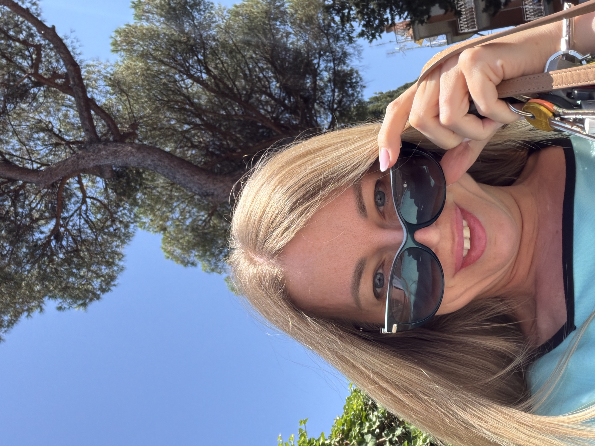 A blonde woman with light blue eyes and pink lipstick smiles at the camera, holding a pair of dark sunglasses in her right hand. She is wearing a teal top with a black trim. Her hair is long and blonde, framing her face. In her right hand, she also holds a brown strap with a keychain containing multiple keys. The background features a large tree with dark green foliage against a bright blue sky. A building with balconies and a light-colored facade is visible in the upper right corner. The scene appears to be outdoors in Rome, Italy.