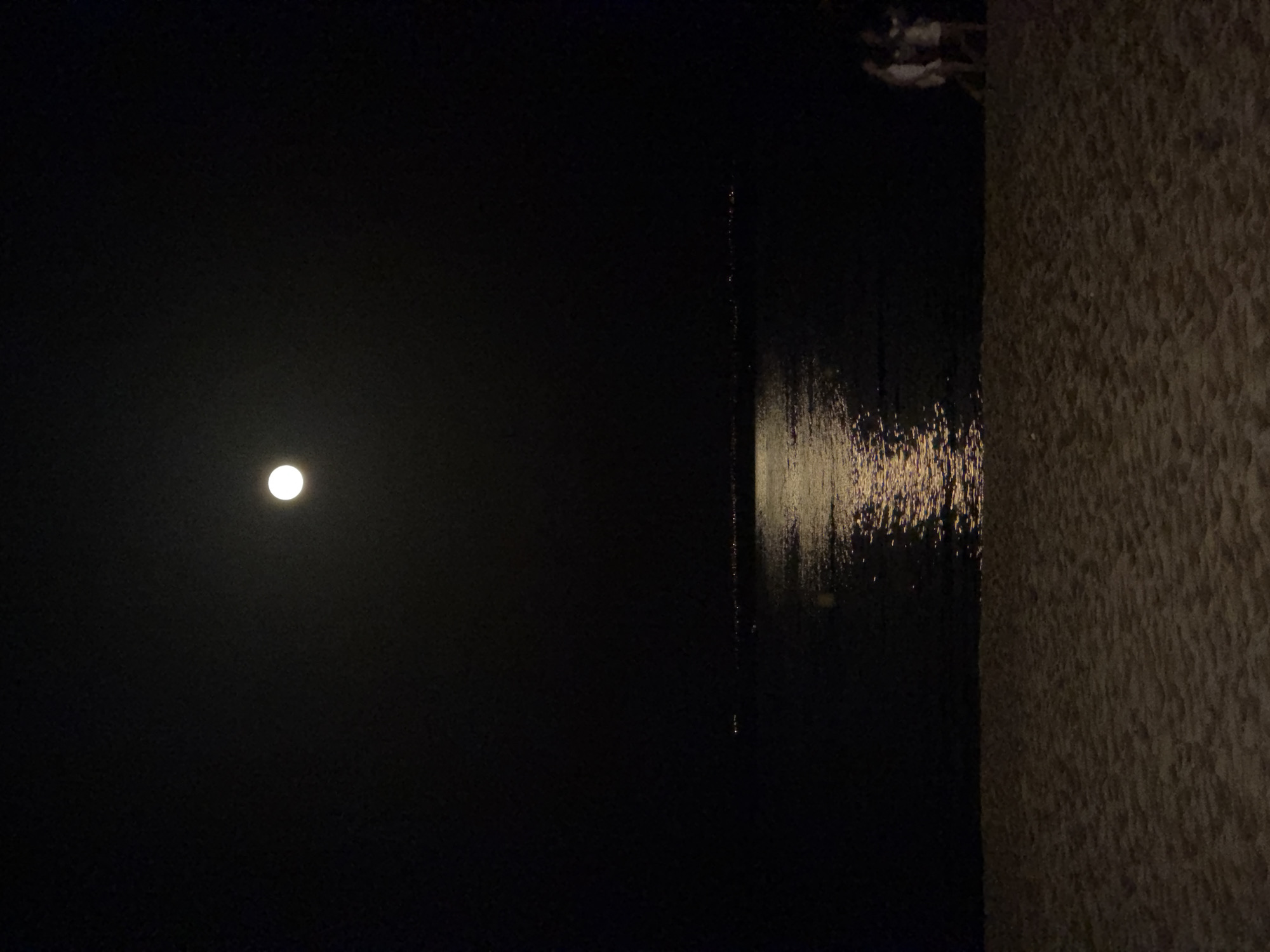 The image depicts a nighttime scene at a beach in Palmanova, Spain. In the upper portion of the frame, a full moon is visible against a dark sky. Below the moon, a body of water reflects the moonlight, creating a shimmering path on the surface. In the distance, along the shoreline, a faint line of lights suggests the presence of buildings or a town. The foreground of the image is occupied by a sandy beach. On the right side of the beach, near the water's edge, three individuals are visible. Two of them appear to be walking together, while the third is slightly behind. They are silhouetted against the dark environment, with their attire appearing light-colored. The overall atmosphere is serene and quiet.