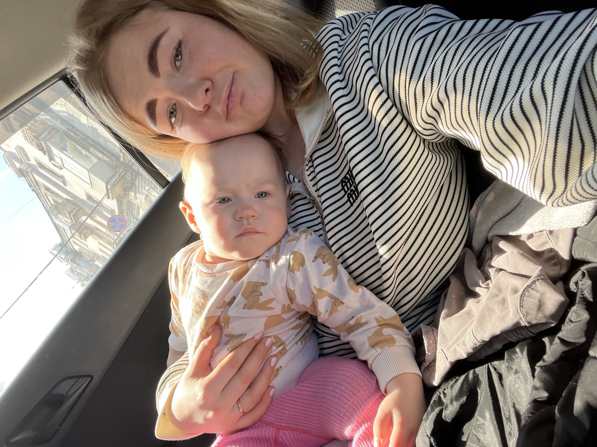A woman holds a baby in her lap inside a vehicle. The woman has fair skin, blonde hair, and a pierced nose, and she wears a striped jacket. The baby has fair skin, blue eyes, and is wearing a patterned shirt and pink pants. The background shows a building visible through the car window, identifiable as being in Perm, Russia, based on the architectural style and street sign. A pile of clothing sits to the right of the woman and baby on the seat.