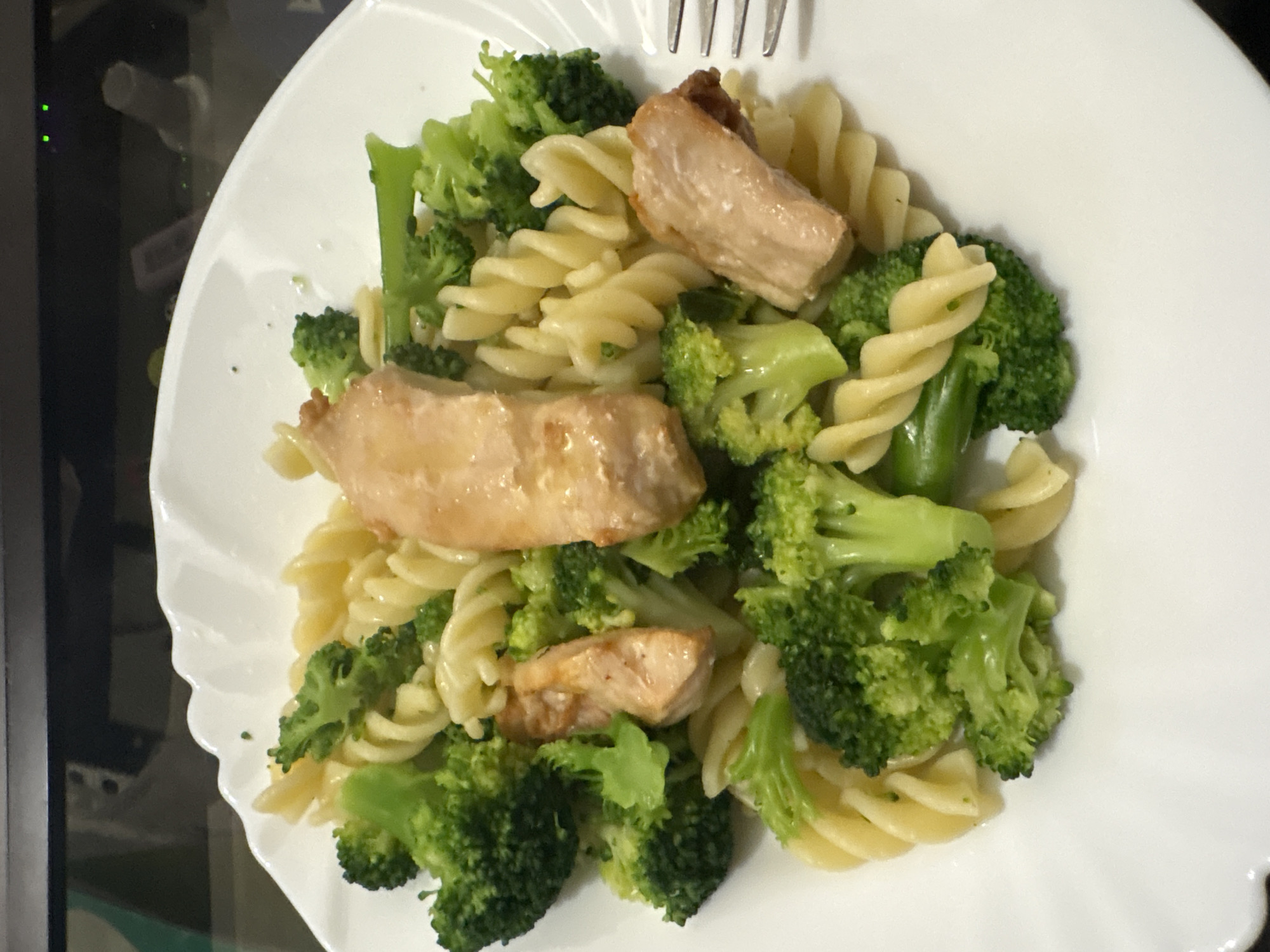 The image shows a close-up, top-down view of a white, fluted plate filled with pasta, broccoli, and pieces of cooked salmon. The pasta is fusilli, and the broccoli florets are bright green and appear to be steamed or lightly sautéed. The salmon pieces are golden brown and appear tender. A fork is partially visible on the right side of the plate. The background is dark and out of focus, suggesting an indoor setting. The image was taken in the city of Bar, Montenegro.