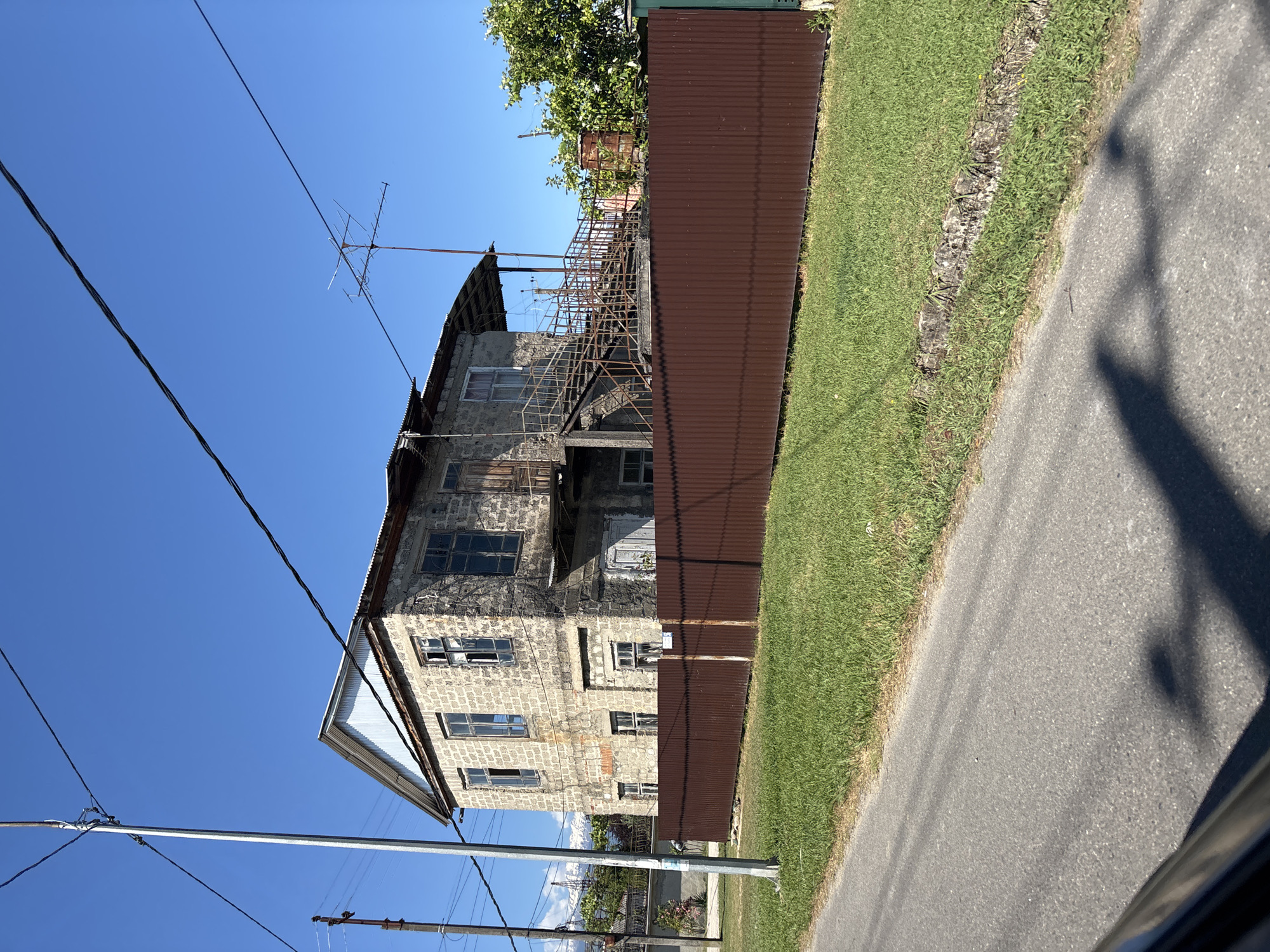 The image depicts a tall, aged, light-colored brick building with a corrugated metal roof. The building appears somewhat dilapidated, with visible external metal stairs on the side, leading up to the upper levels. Several windows are present, some of which appear broken or boarded up. A television antenna is mounted on the roof. The building is situated next to a rusty brown corrugated metal fence, which separates it from a neatly trimmed green lawn. Beyond the fence and the building, there are some trees and what appears to be a clear blue sky. A paved road runs alongside the lawn, and a shadow of a person or object is cast on the road. The scene is likely outdoors during the daytime, given the bright sunlight and the clear sky. The overall impression is one of a rural or semi-rural setting with older architecture. No people are visible in the frame, and no text is discernible.