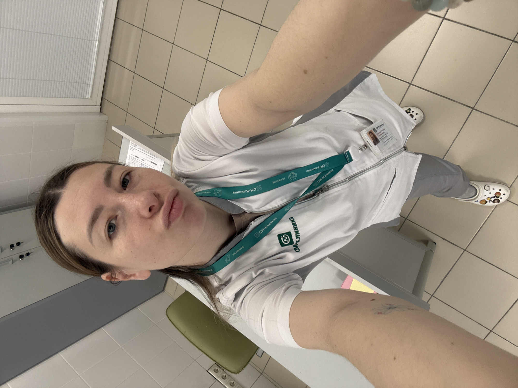 The image is a selfie of a woman indoors. She has fair skin, light brown hair with darker roots, and blue eyes. She is wearing a white medical jacket over a white t-shirt with gray pants and Crocs shoes. A lanyard around her neck has the logo "CM-Klinika." The CM-Klinika logo suggests this is a branch of this medical clinic located in Moscow, Russia. She is standing in a room with tiled flooring. The walls are tiled, with gray and white cabinets. A green chair is also visible. Her facial expression is one of a duck-face pout. She is holding her arm out at an angle, taking the photo. A tattoo can be seen on her left arm.