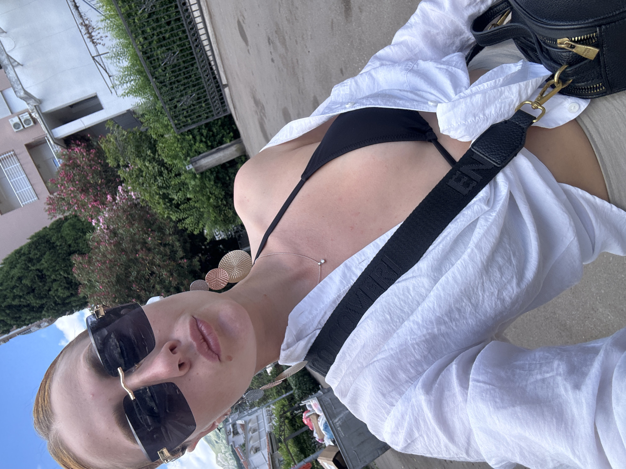 A woman stands outdoors in what appears to be an urban environment. She is wearing large, dark sunglasses, a black bikini top, and an unbuttoned white shirt. She has voluminous blonde hair pulled back. Dangling earrings, a delicate necklace, and earbuds are visible. She carries a black bag with a prominent strap across her chest. In the background, buildings, trees, and a large metal dumpster are visible. The scene suggests a warm day, possibly in a Mediterranean setting, given the architecture and vegetation. The ground is paved, indicating a street or walkway. The lighting suggests it is daytime, with the sun casting shadows. The woman's pose suggests she is taking a selfie.