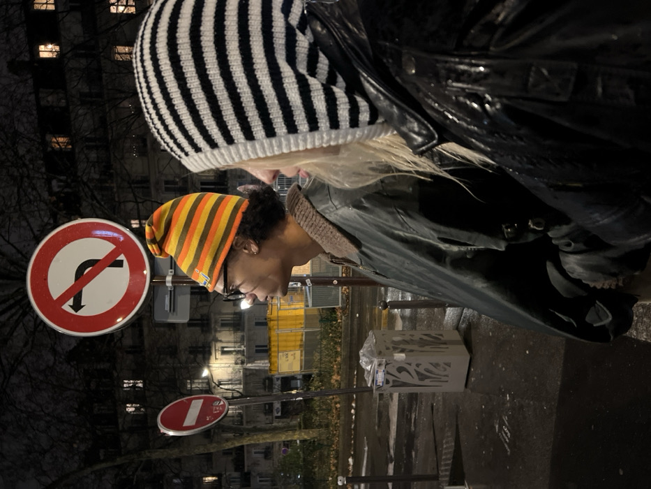 Two individuals stand near street signs and buildings at night. One person wears a black and white striped beanie and a black jacket. The other wears a beanie with orange, yellow, and brown stripes and glasses. Both appear to be looking downward. A “no turning” street sign is prominently displayed, along with another sign showing a white bar in a red circle. A decorative trash receptacle is visible. Buildings with lit windows line the street in the background.