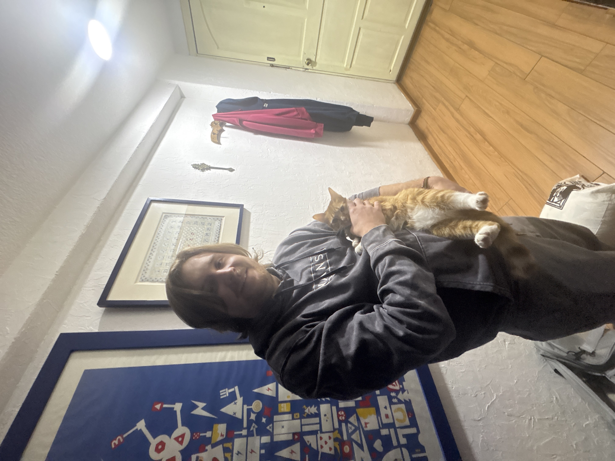 A person with long hair, wearing a gray sweatshirt, is holding an orange tabby cat. The person is positioned in front of a wall with two framed pictures. Behind them are a white door, a coat rack with clothing, and a wooden floor. The location appears to be inside a home. There is no information in the image to indicate that the location is in Mexico City, Mexico.