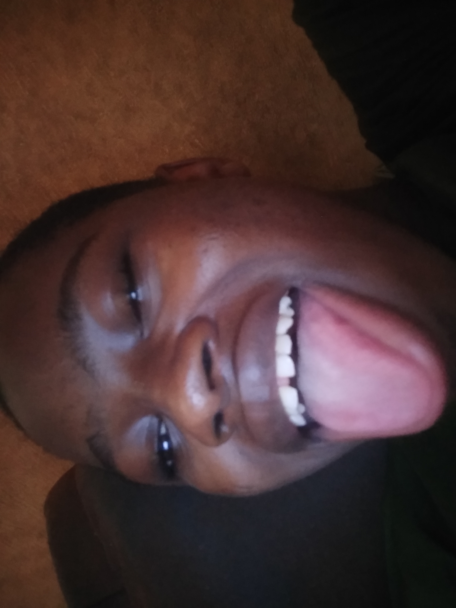 A close-up, tilted perspective photo depicts a person with dark skin protruding their tongue. The person's eyes are open, but unfocused. The background shows a blurred mix of dark and light brown surfaces. No specifics related to Shangisha, Nigeria, or any other location can be ascertained from the image.