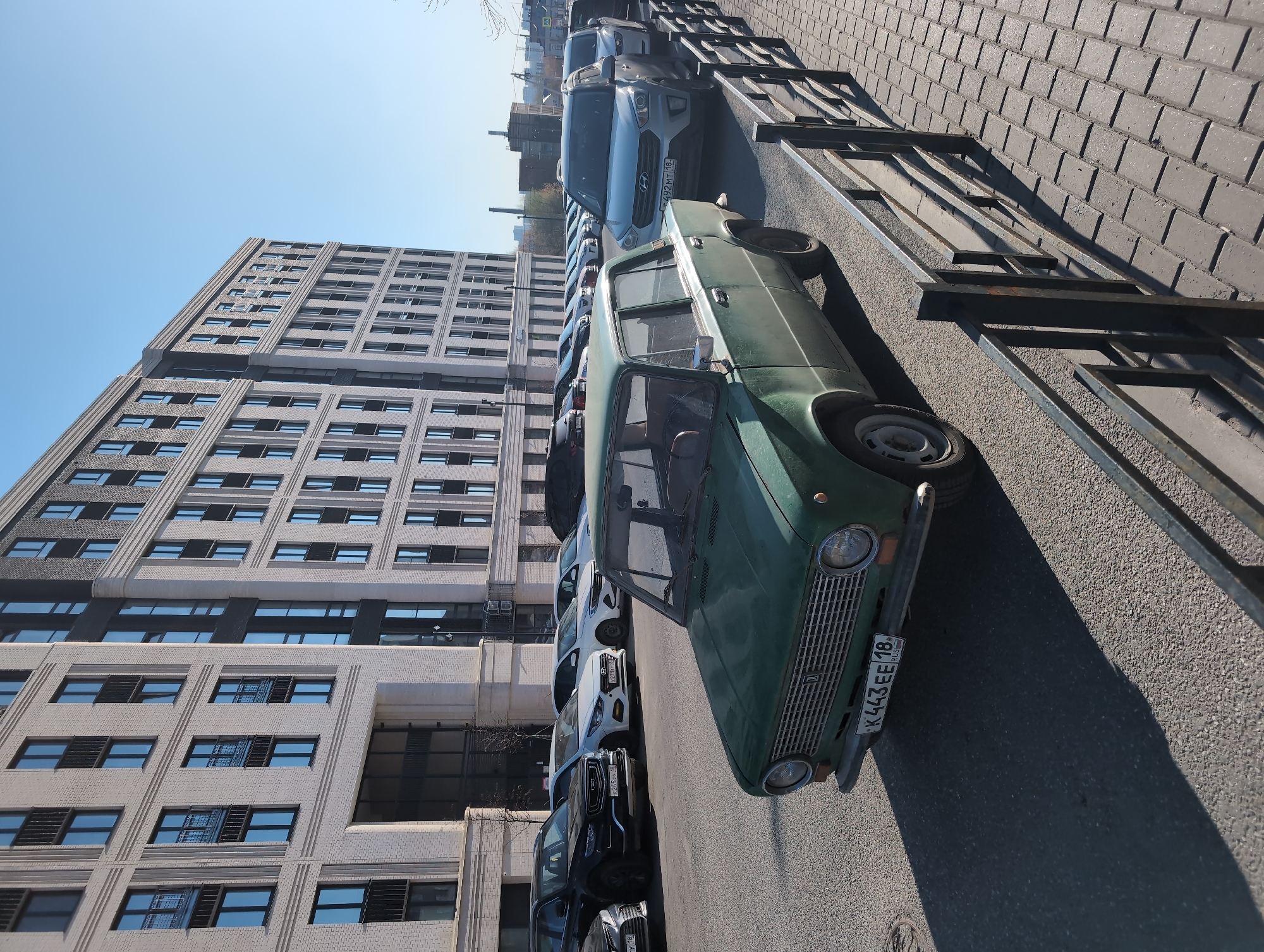 The image shows a street scene in Izhevsk, Russia. A green Lada car is parked on the right side of the street, next to a black metal fence separating the sidewalk from the road. A row of modern cars is parked further down the street. In the background, a large, modern building with many windows rises several stories high. The sky is clear and blue. The license plate on the Lada reads "K443EE 18 RUS." The sidewalk is paved with gray brick. The road surface is asphalt. No people are visible in the image.