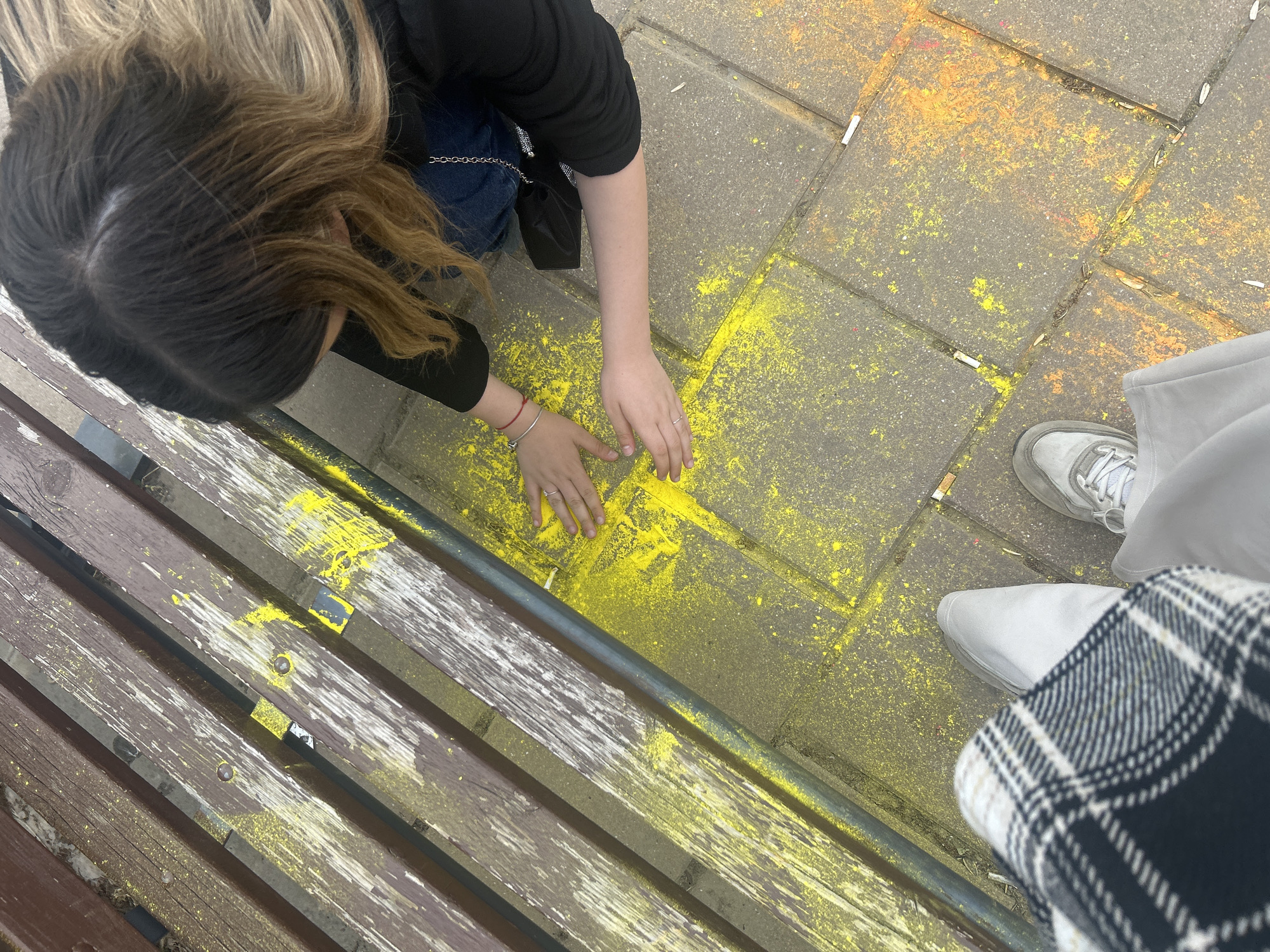 A high-angle shot captures a scene on a stone-tiled surface, likely a public space, near a wooden bench. A person with long brown and blonde hair, wearing a black top and jeans, kneels and places their hands on the ground covered with yellow powder. Another person stands nearby, wearing light-colored shoes and pants with a black and white checkered pattern. Cigarette butts are scattered on the ground, mixed with yellow and orange powder. The bench is weathered with visible paint chipping and traces of the yellow powder. This appears to be located in the city of Ruse, Bulgaria.