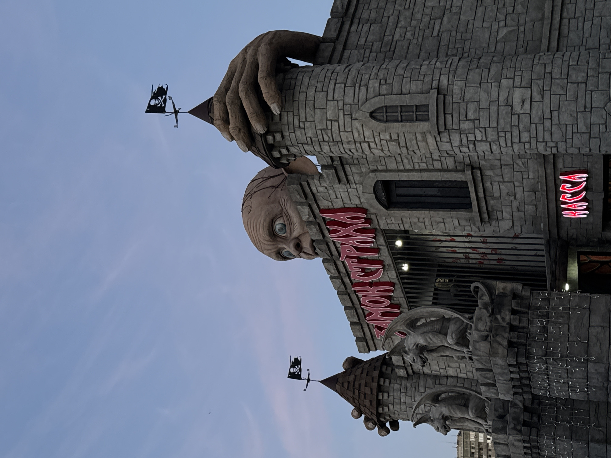 The image shows the facade of a building in Yevpatoriya, Ukraine, specifically the "Castle of Horrors". The building is designed to resemble a medieval stone castle with multiple towers and turrets. A large, grotesque head with bulging eyes and red, bloody streaks extending down from its mouth is visible on the facade. An oversized hand appears to grasp the building from above. There are gargoyle statues on the lower parts of the building. Pirate flags are placed on top of the towers. The sky is visible in the background, indicating it is either dusk or dawn. Neon signs in Cyrillic script are mounted on the building.